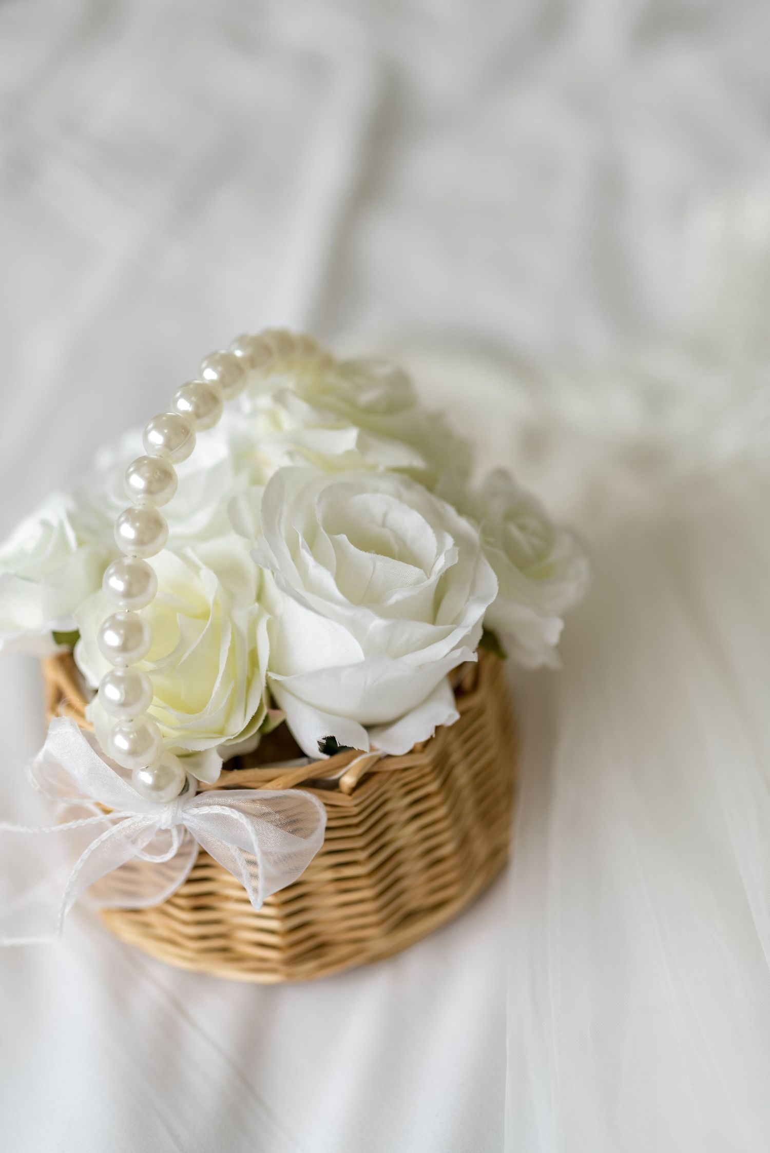 Wicker basket filled with white roses and a pearl necklace tied with a sheer bow on white fabric.