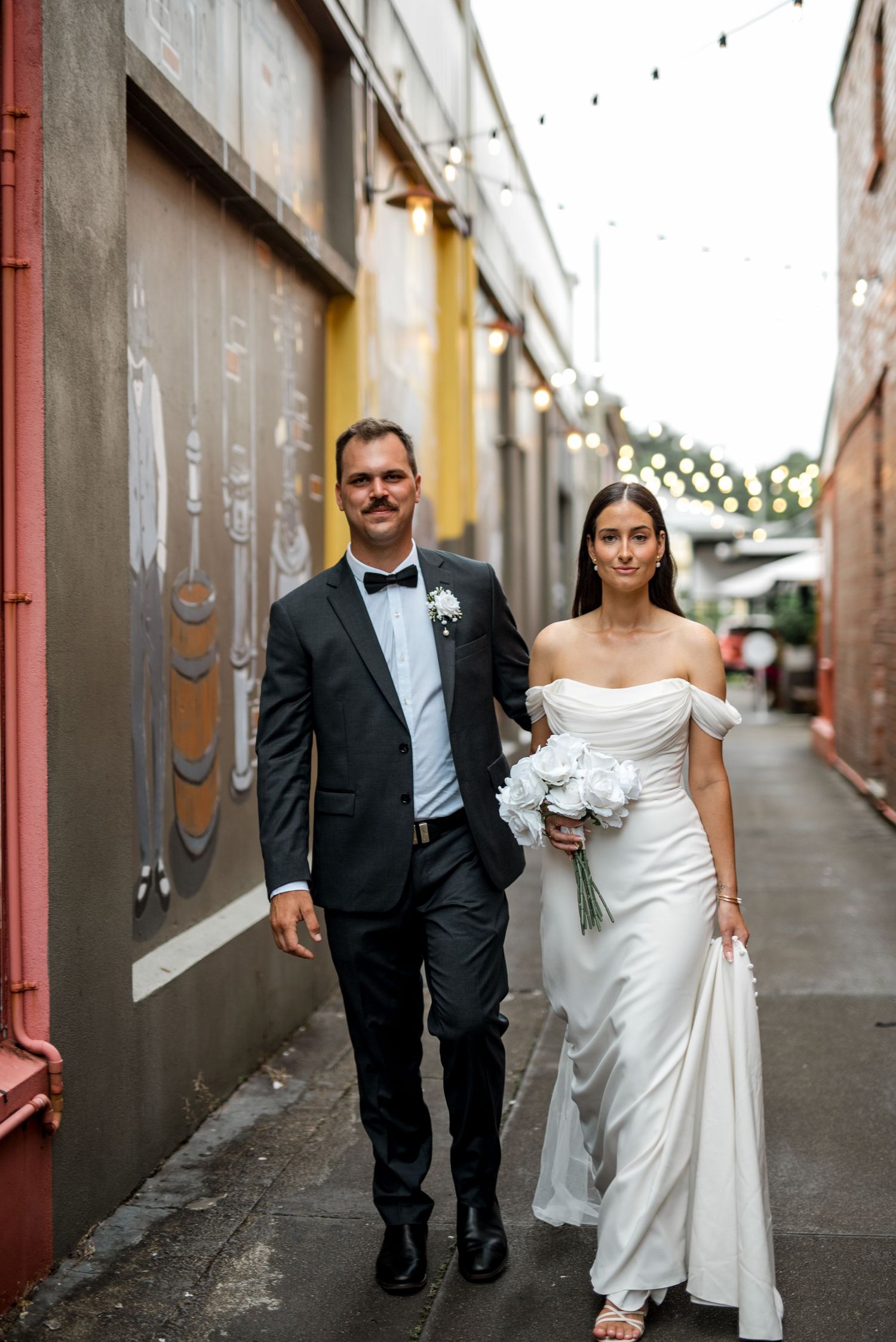 Elegant bride and groom walk hand in hand through a laneway with string lights above.