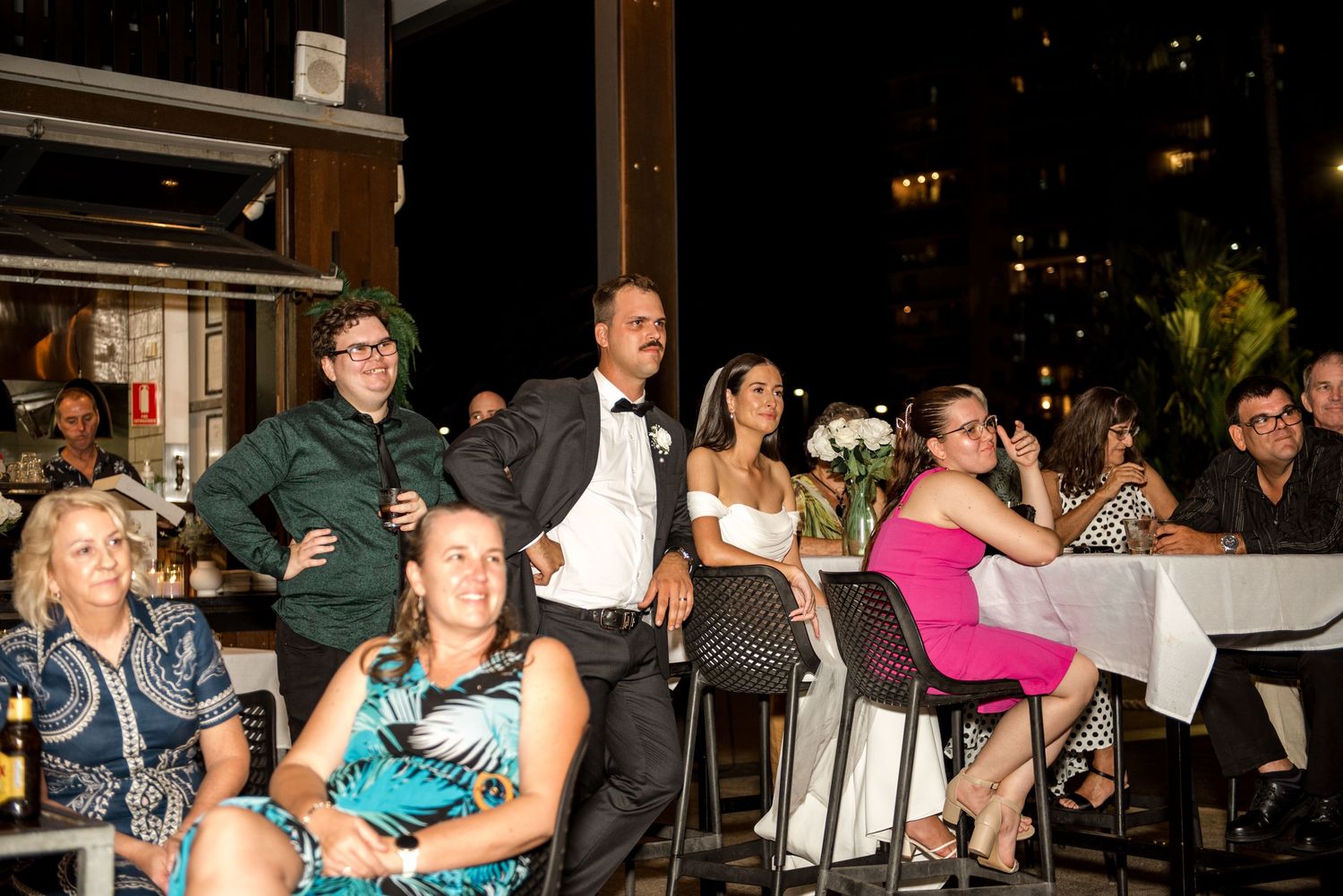 Wedding guests watch speeches at an outdoor evening reception, with bride in white and groom in tuxedo seated among attendees.