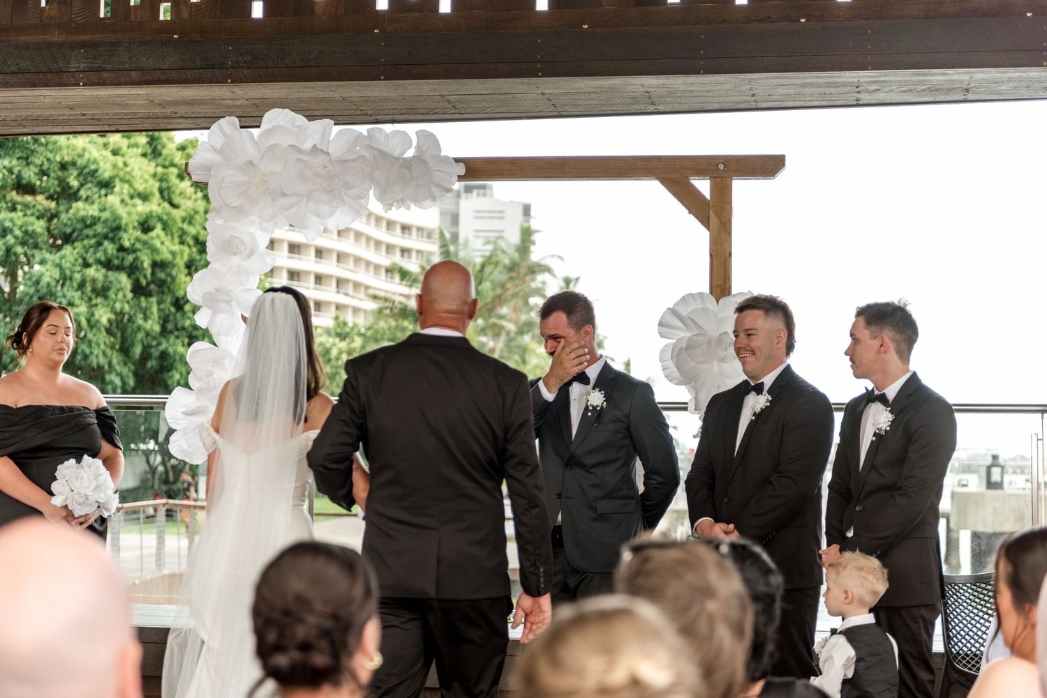 Groom wipes tears as bride approaches altar at outdoor waterfront wedding ceremony.