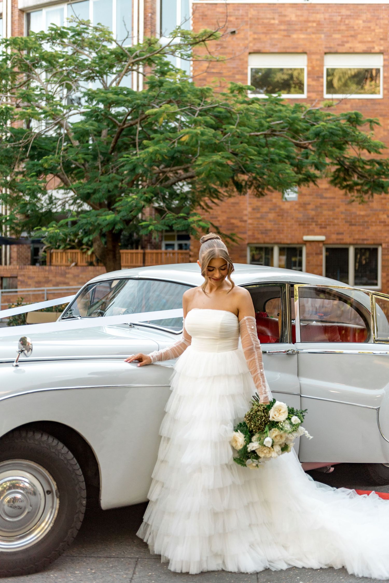 Bride in strapless tiered gown poses beside vintage white classic wedding car.