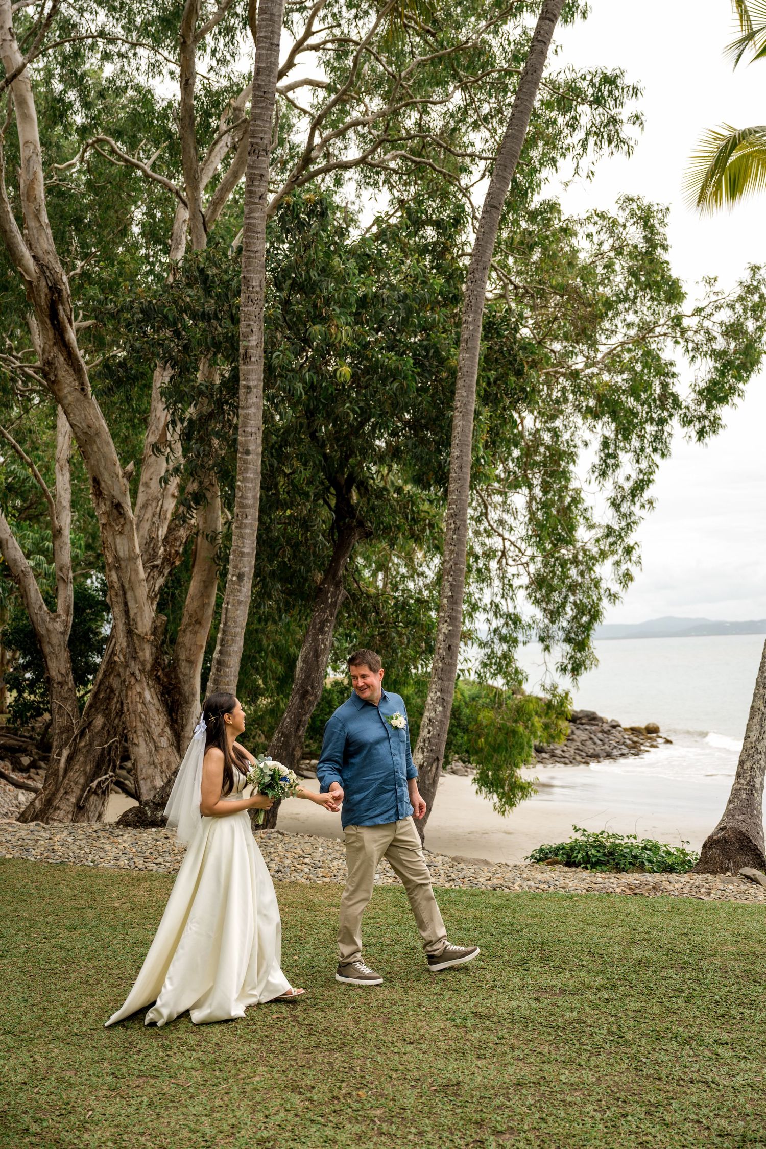 Bride and groom walk together on a tropical beachside lawn surrounded by tall trees and ocean views at Little Cove, Port Douglas.