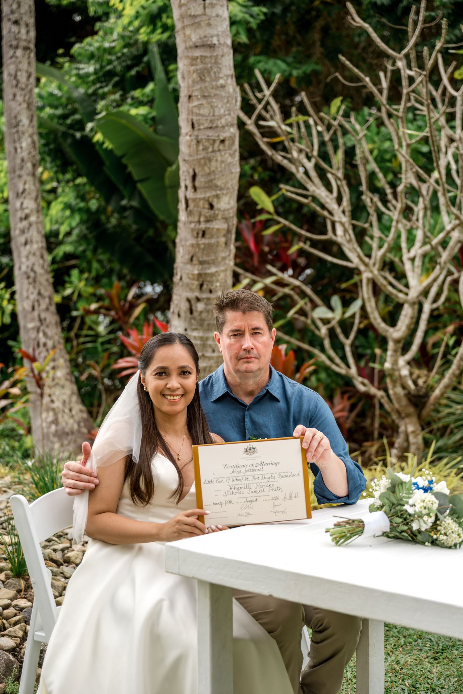 Smiling newlyweds display official marriage certificate at white table in tropical garden at Little Cove, Port Douglas.