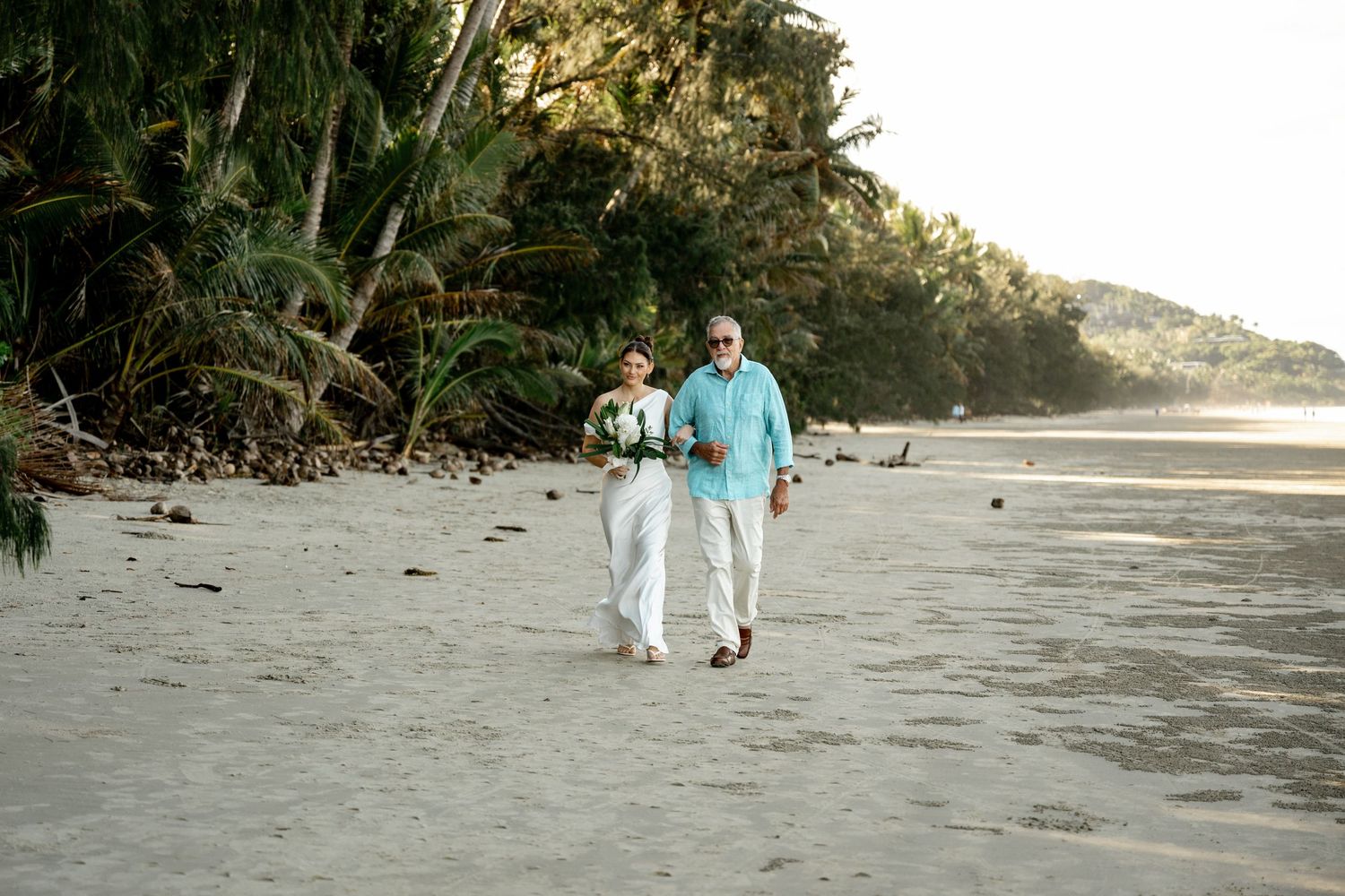 Couple walking hand in hand on a tropical beach, bride holding white bouquet, lush palm trees in background at Four Mile Beach.