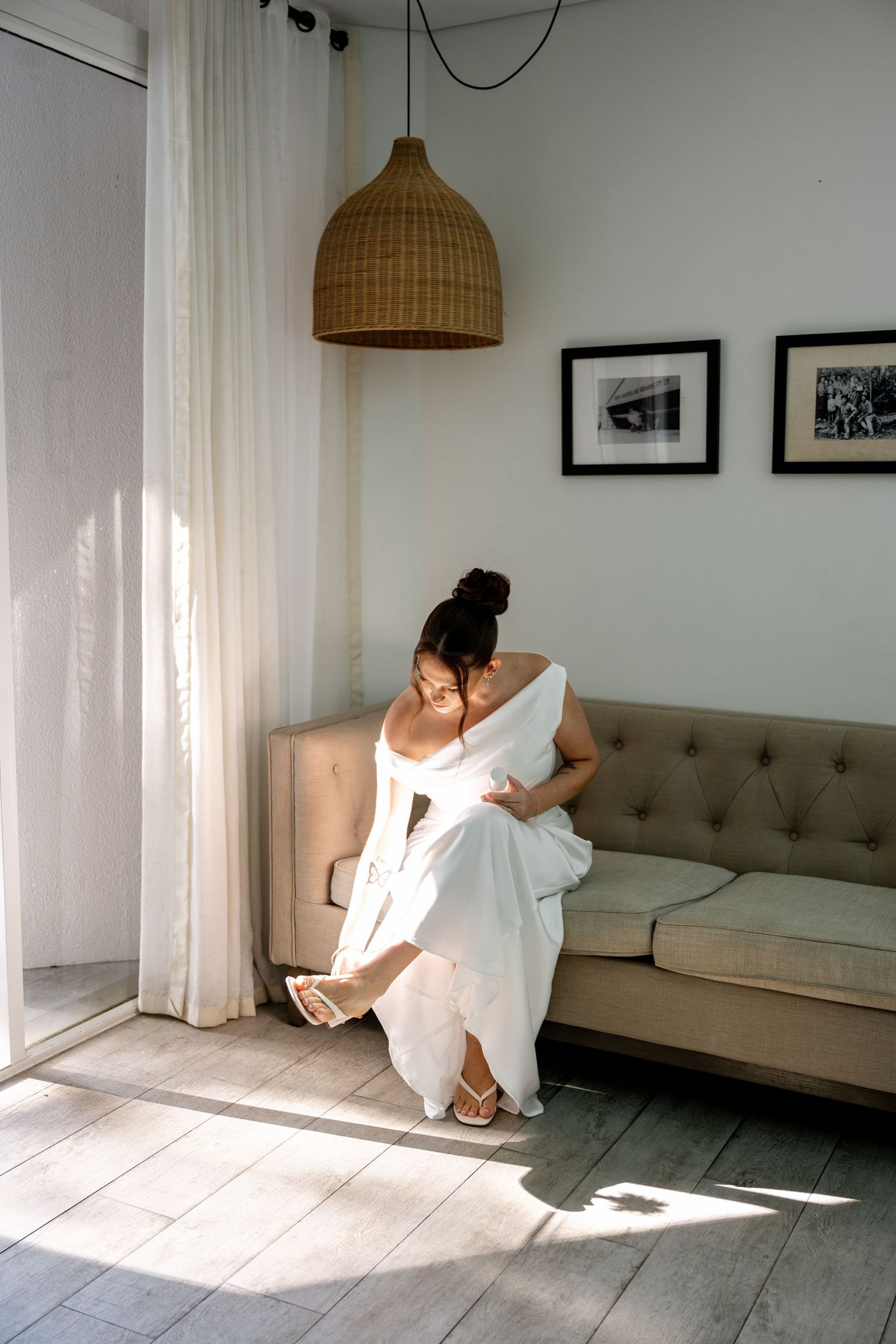 Bride in elegant white wedding gown sits on beige sofa putting on shoes in sunlit modern room.