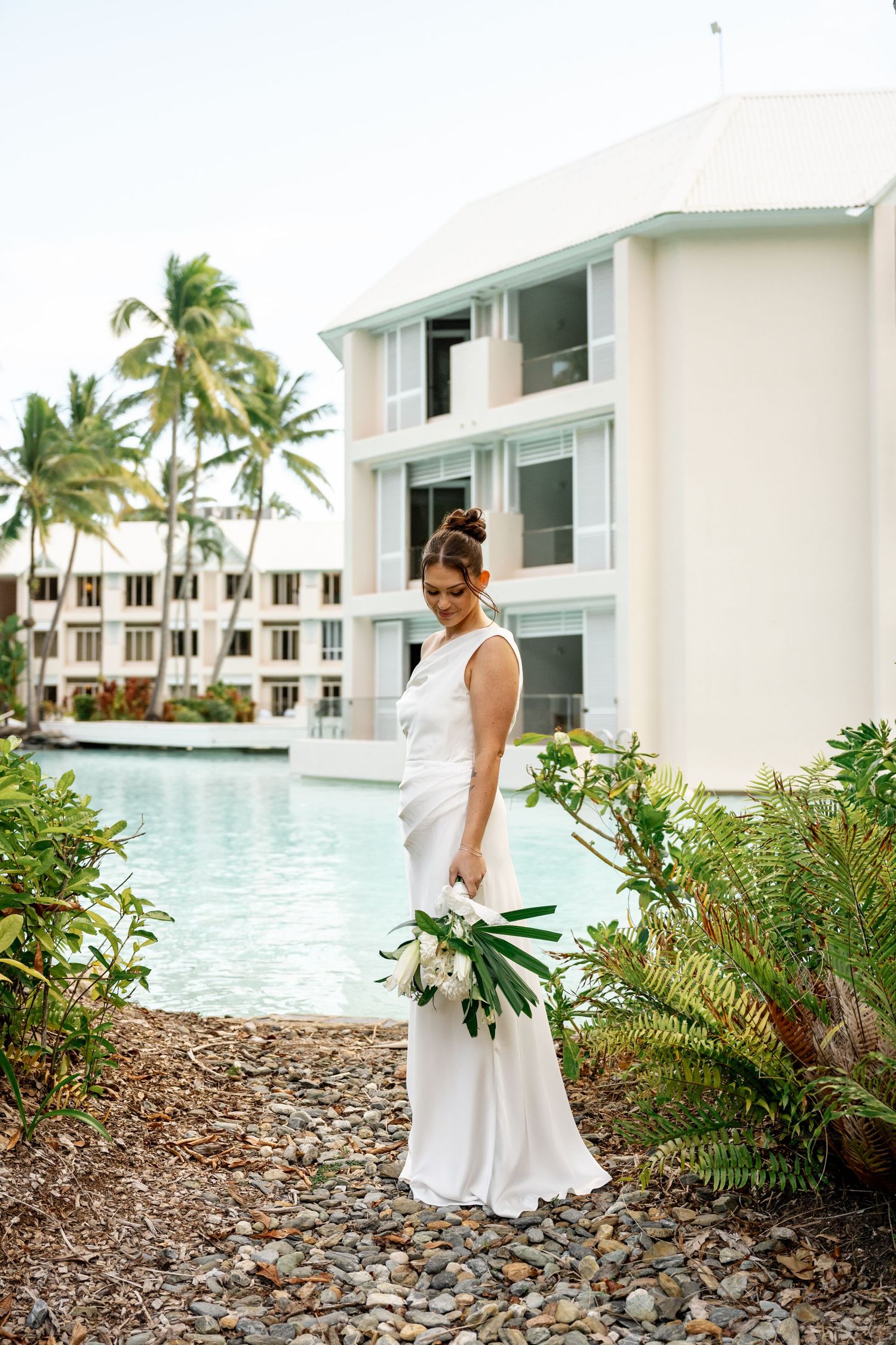 Bride in white gown looking down at her tropical bouquet beside a luxury resort swimming pool at Sheraton Grand Mirage Port Douglas.