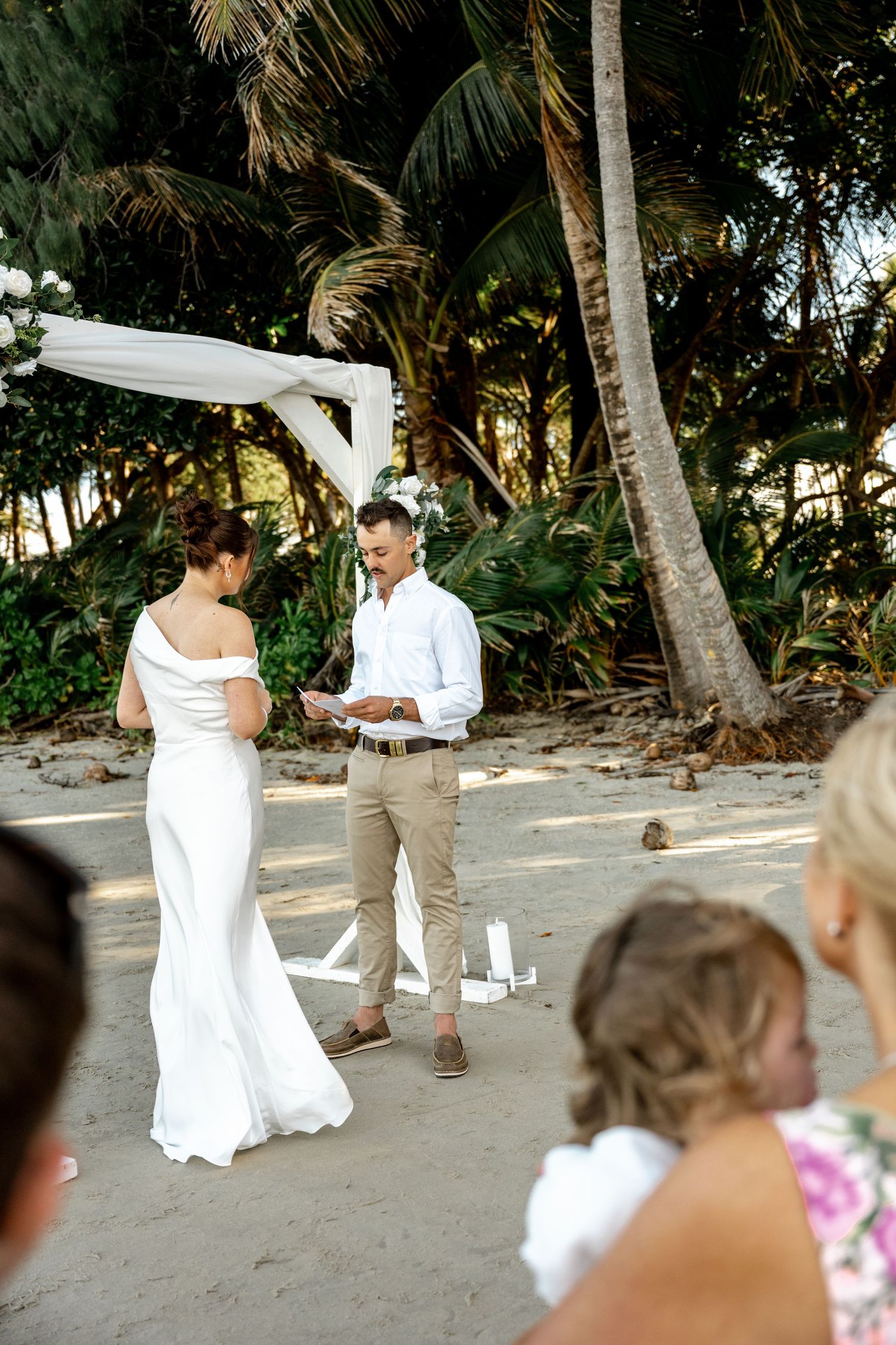 Groom reading wedding vows to bride under a draped white arch on a sandy beach surrounded by palm trees at Four Mile Beach, Port Douglas.