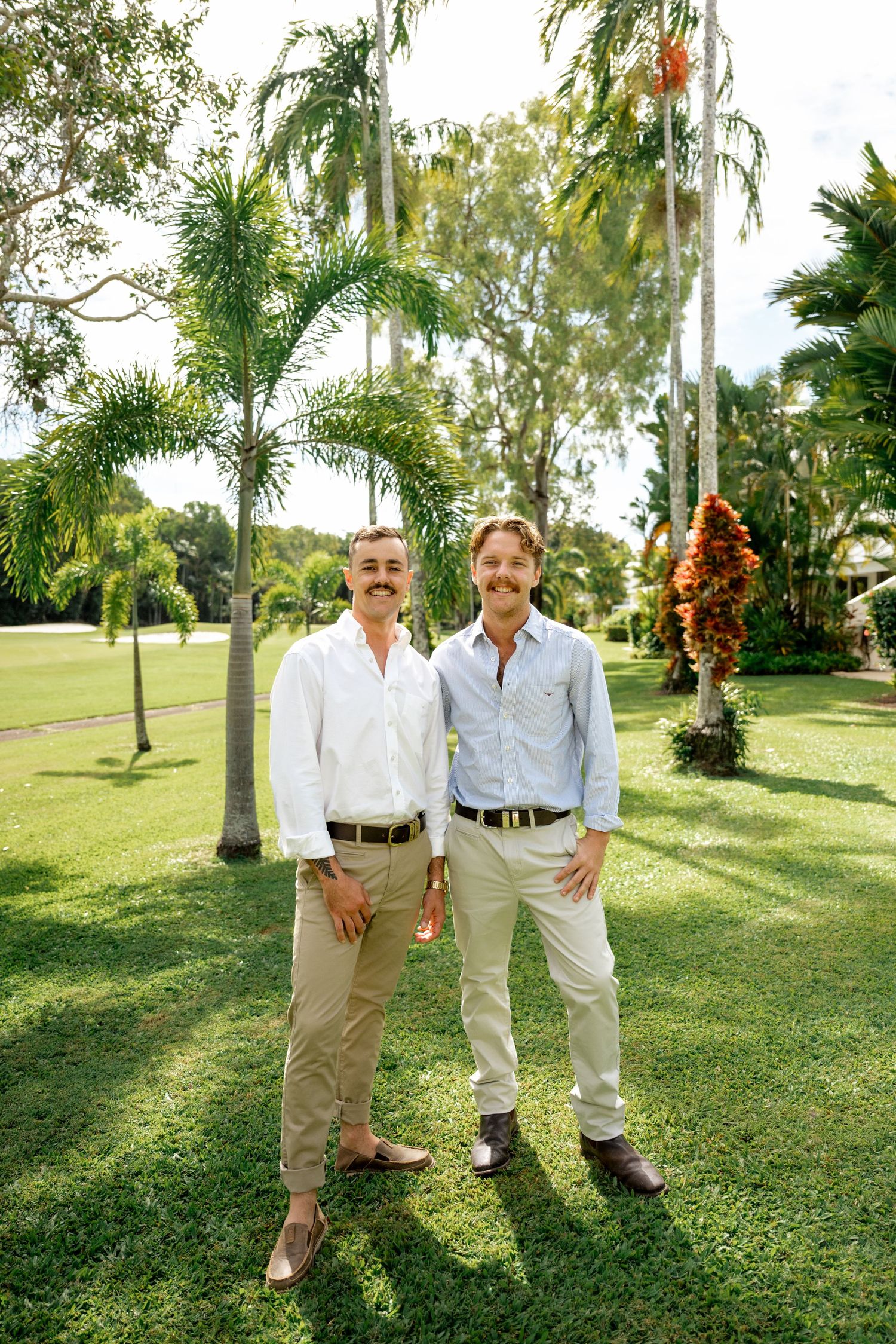 Two men with mustaches in light shirts smiling together in a lush tropical garden setting.