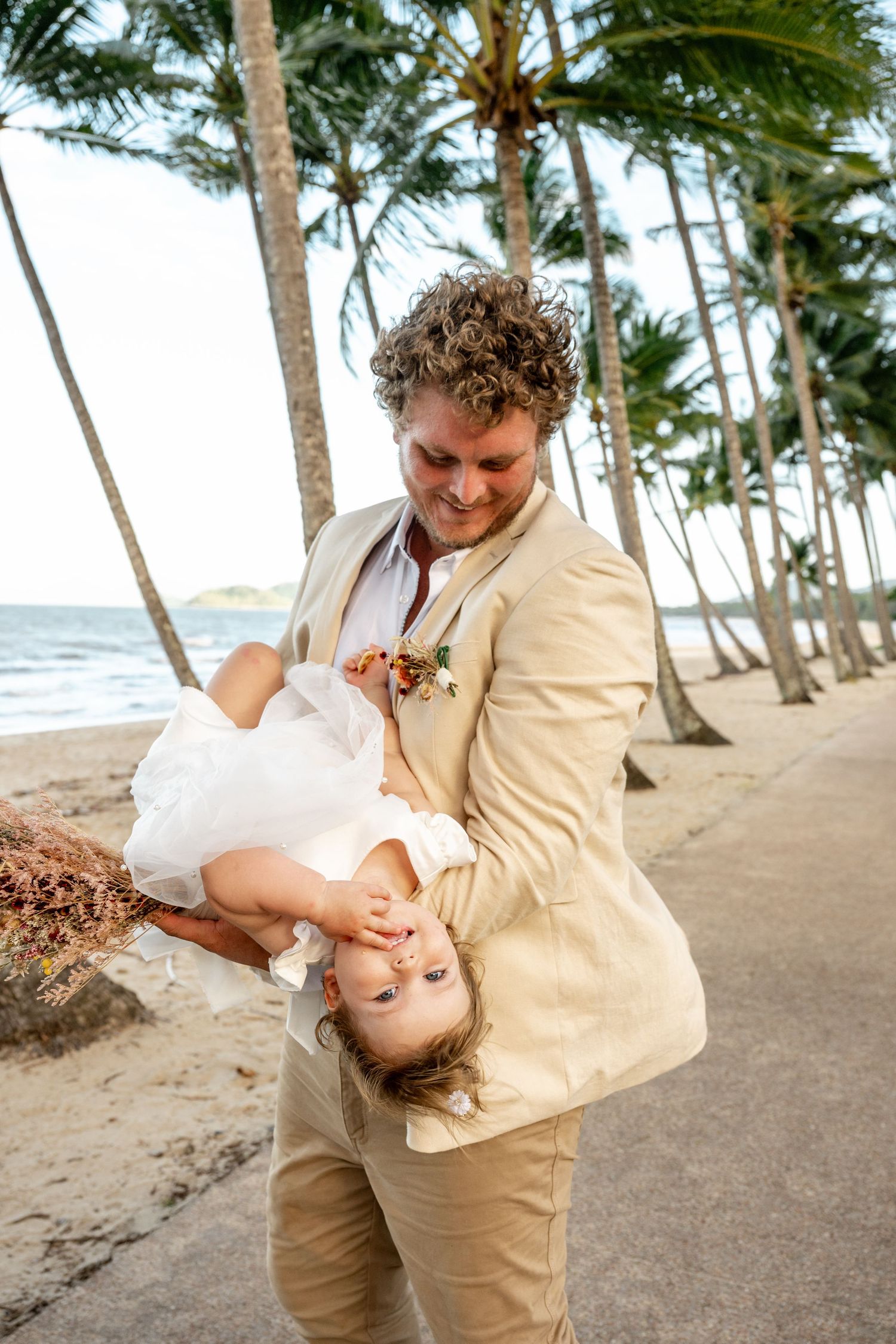 Groom playfully dipping and spinning bride upside down on a tropical beachside path lined with palm trees.