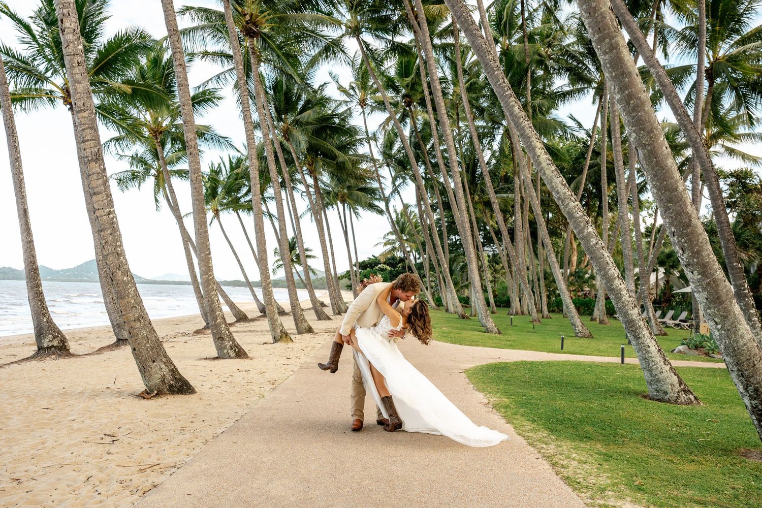 Wedding couple sharing a dip kiss among tall palm trees on a sandy tropical beach pathway.