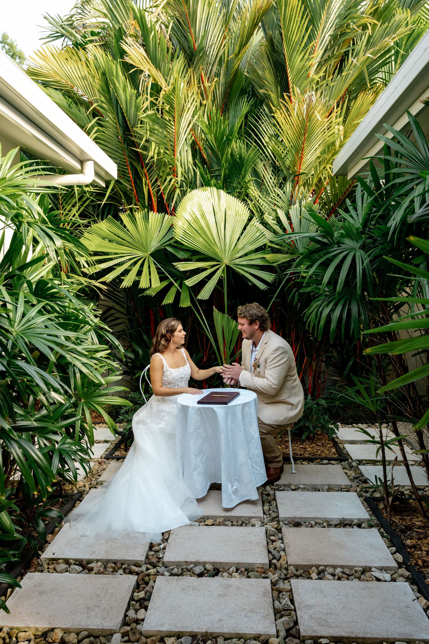 Bride and groom sit together at signing table surrounded by lush tropical palm trees.