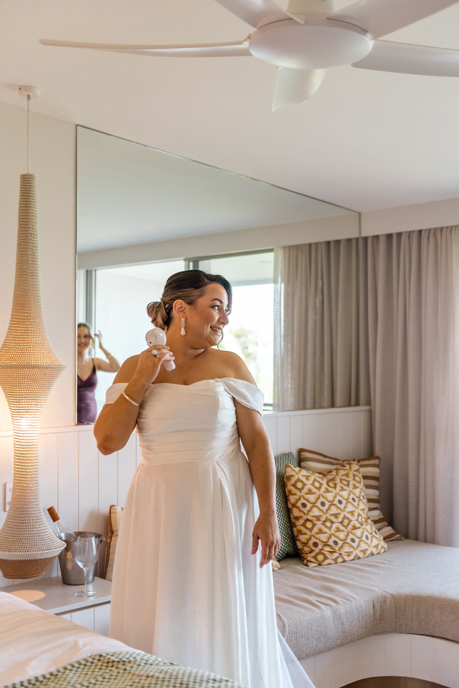 Smiling bride in white off-shoulder wedding dress putting on earrings in bright resort hotel room.