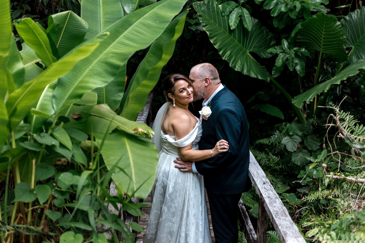 Bride and groom embrace romantically among lush tropical greenery on their wedding day.