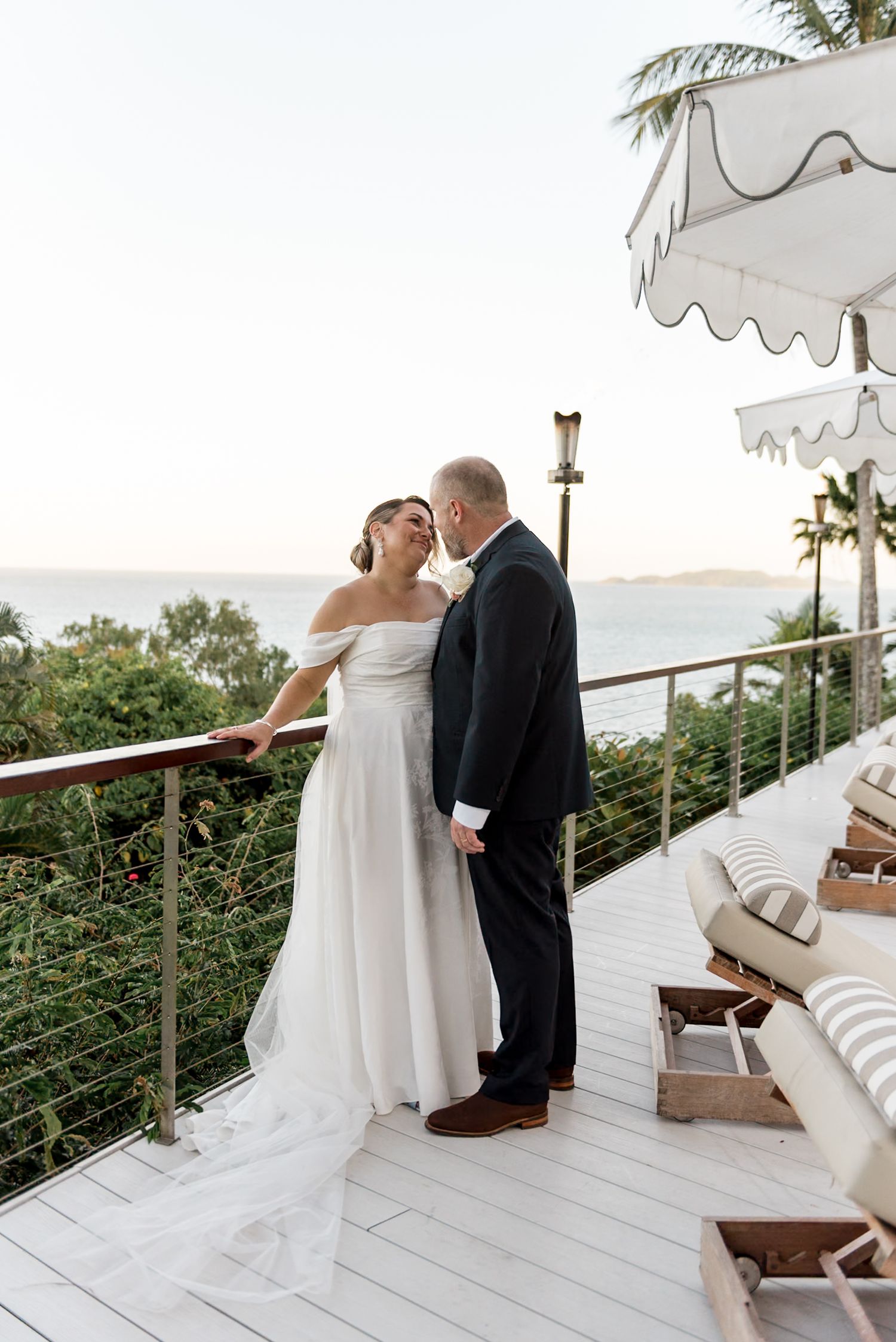 Bride and groom standing close together on a white deck with stunning ocean sunset views.