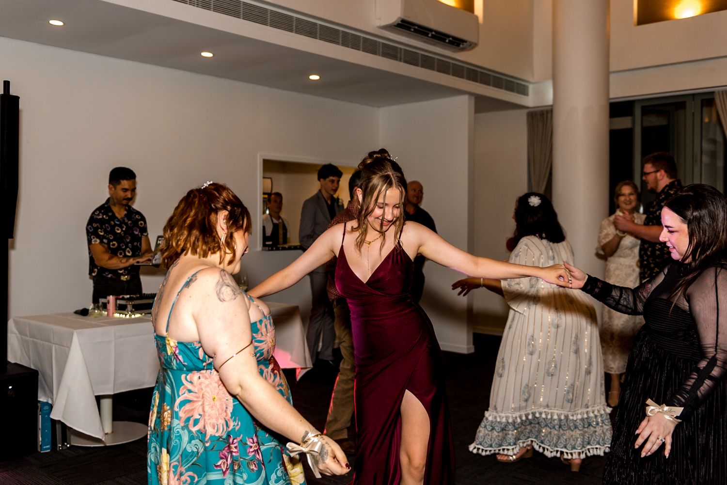 Three women in colorful dresses dance together at a lively wedding reception.