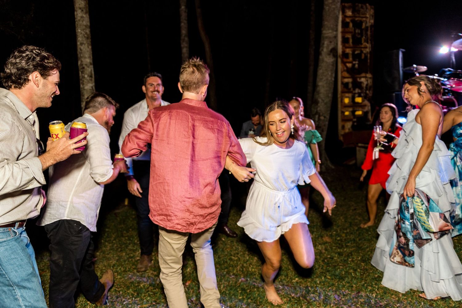 Guests dancing joyfully at an outdoor wedding reception at night, with string lights and tropical trees in the background.