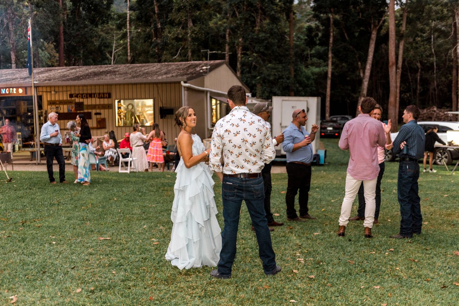 Bride in white gown dancing with groom on grass lawn while wedding guests socialize in background at outdoor reception.