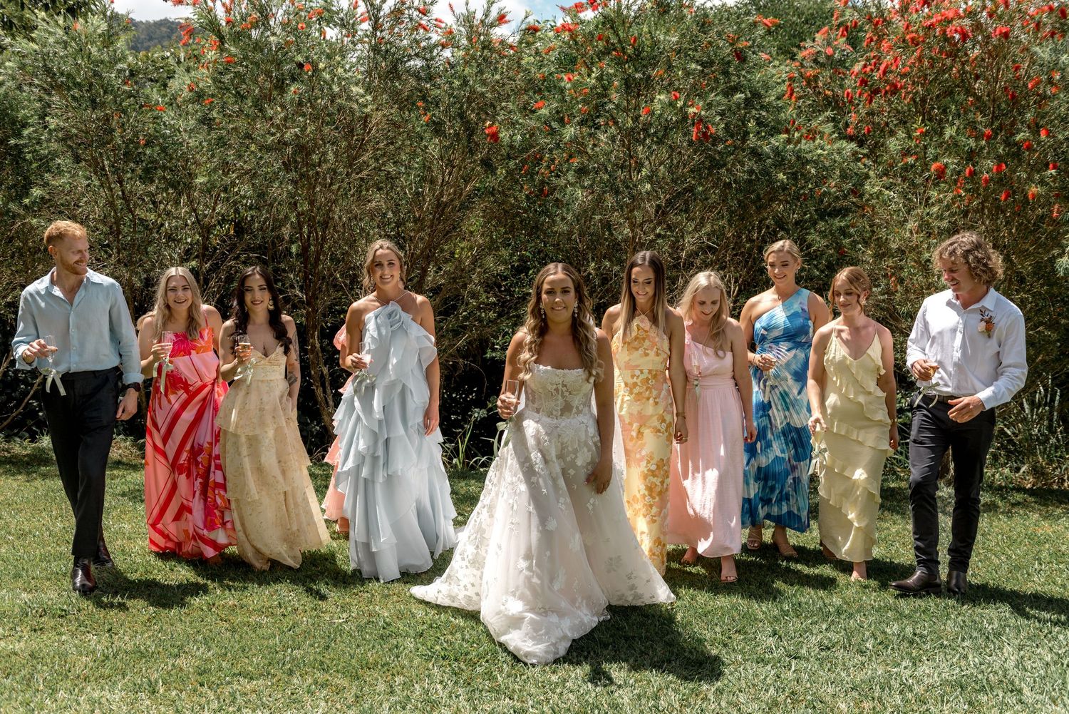 Bride with bouquet poses alongside wedding party in colorful dresses before blooming garden.
