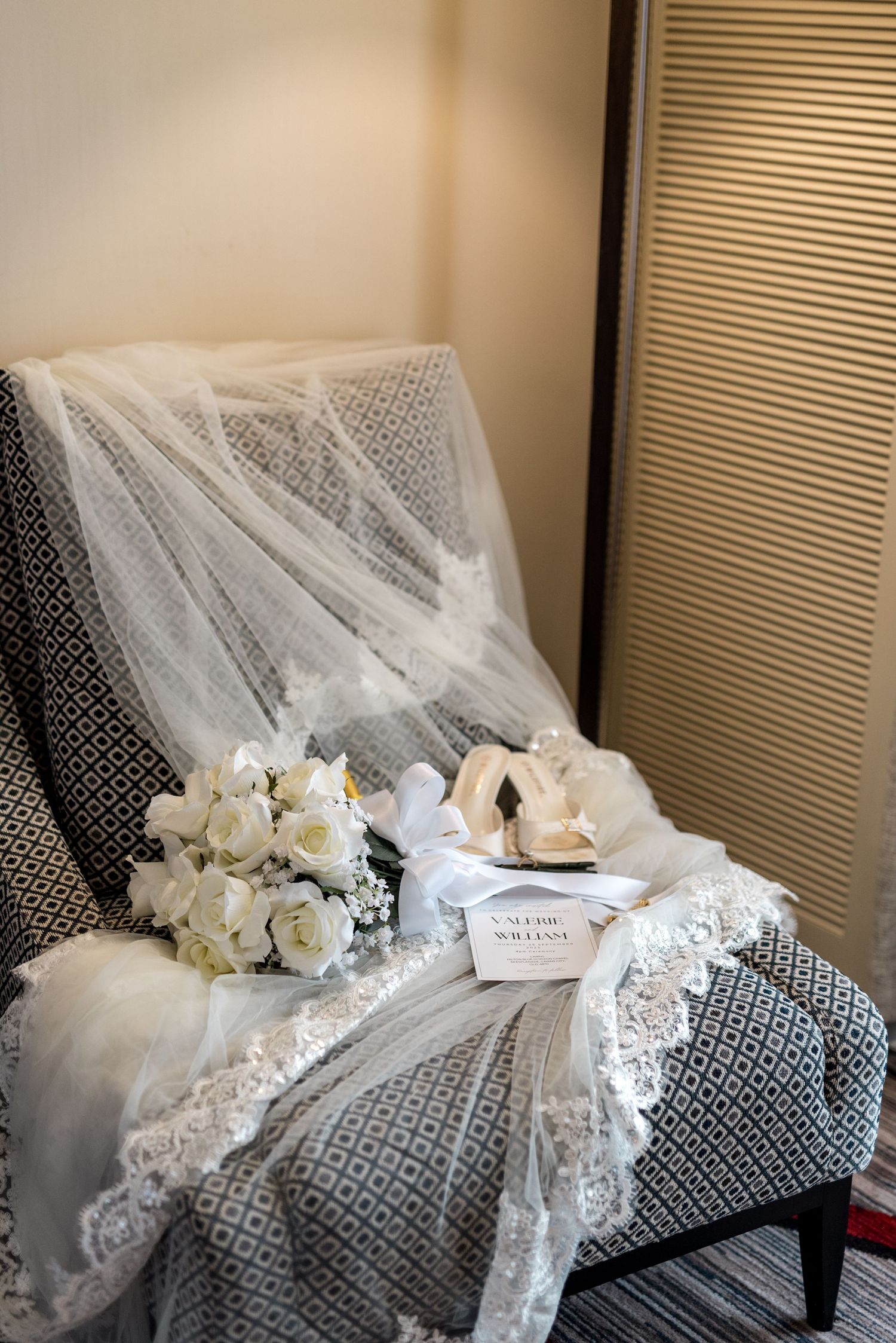 Bridal accessories arranged on a patterned chair including white rose bouquet, veil, heels, and wedding invitation.