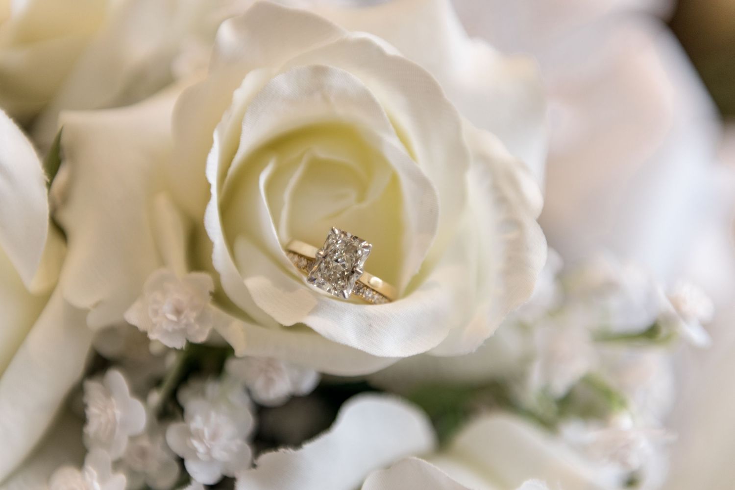 Diamond engagement ring nestled inside a white rose surrounded by baby's breath in a bridal bouquet.