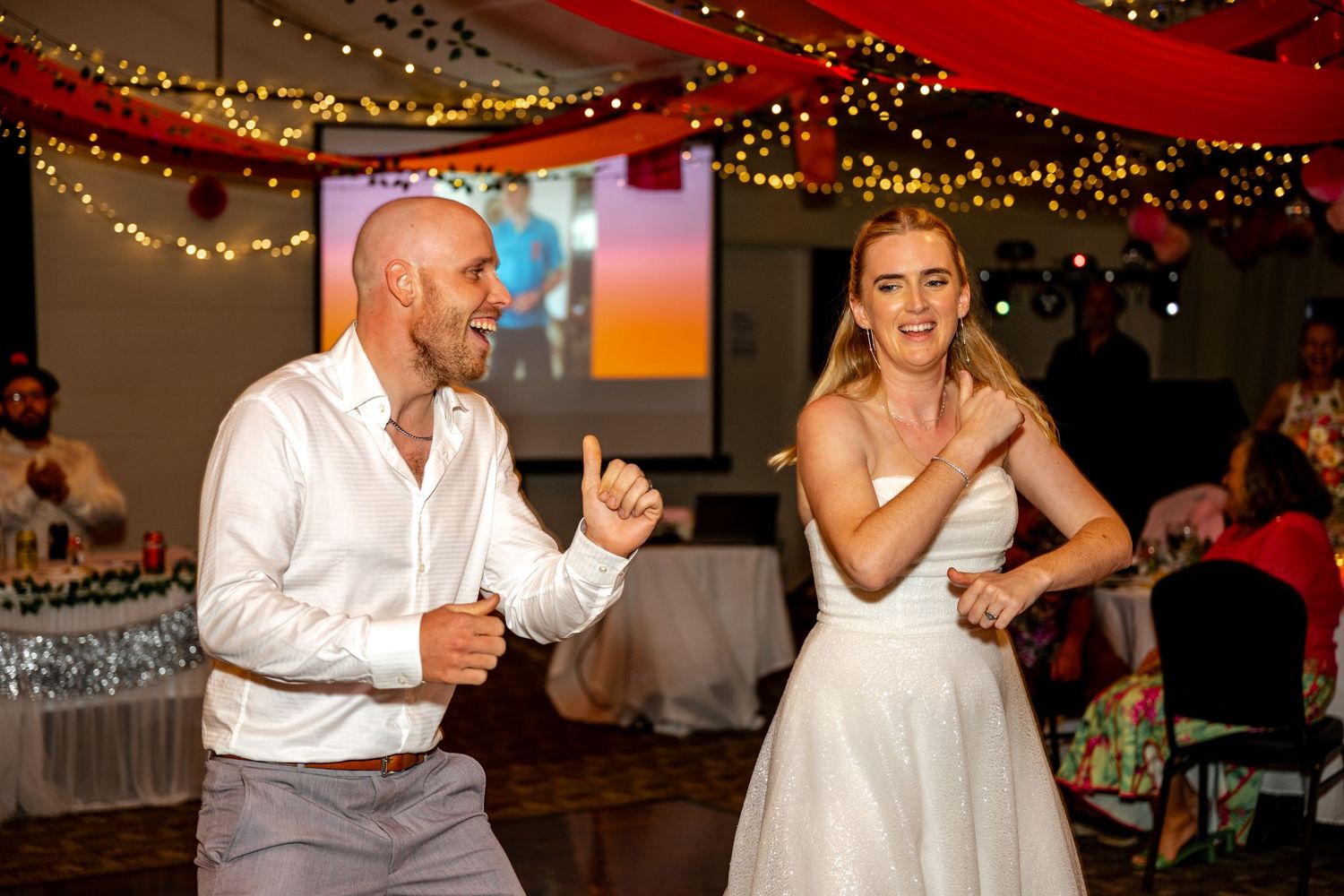 Bride and groom laugh together while dancing at a lively wedding reception with guests watching.