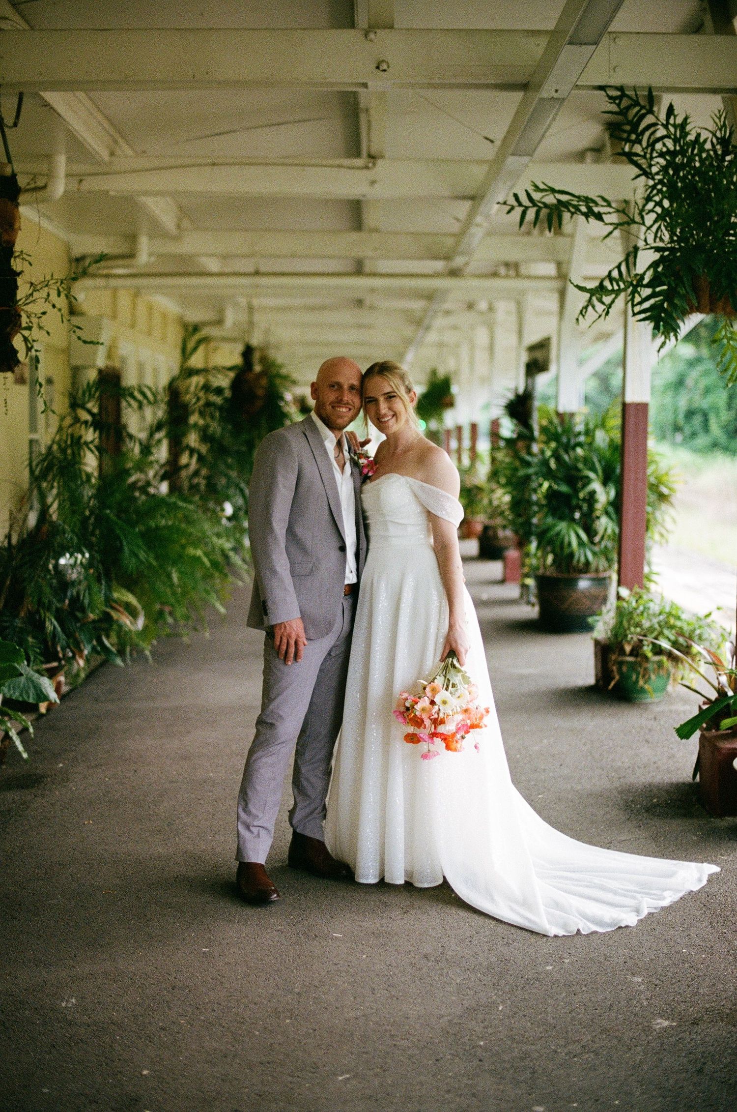 Bride and groom posing in a plant-lined covered walkway, bride holding a coral and pink tropical bouquet.