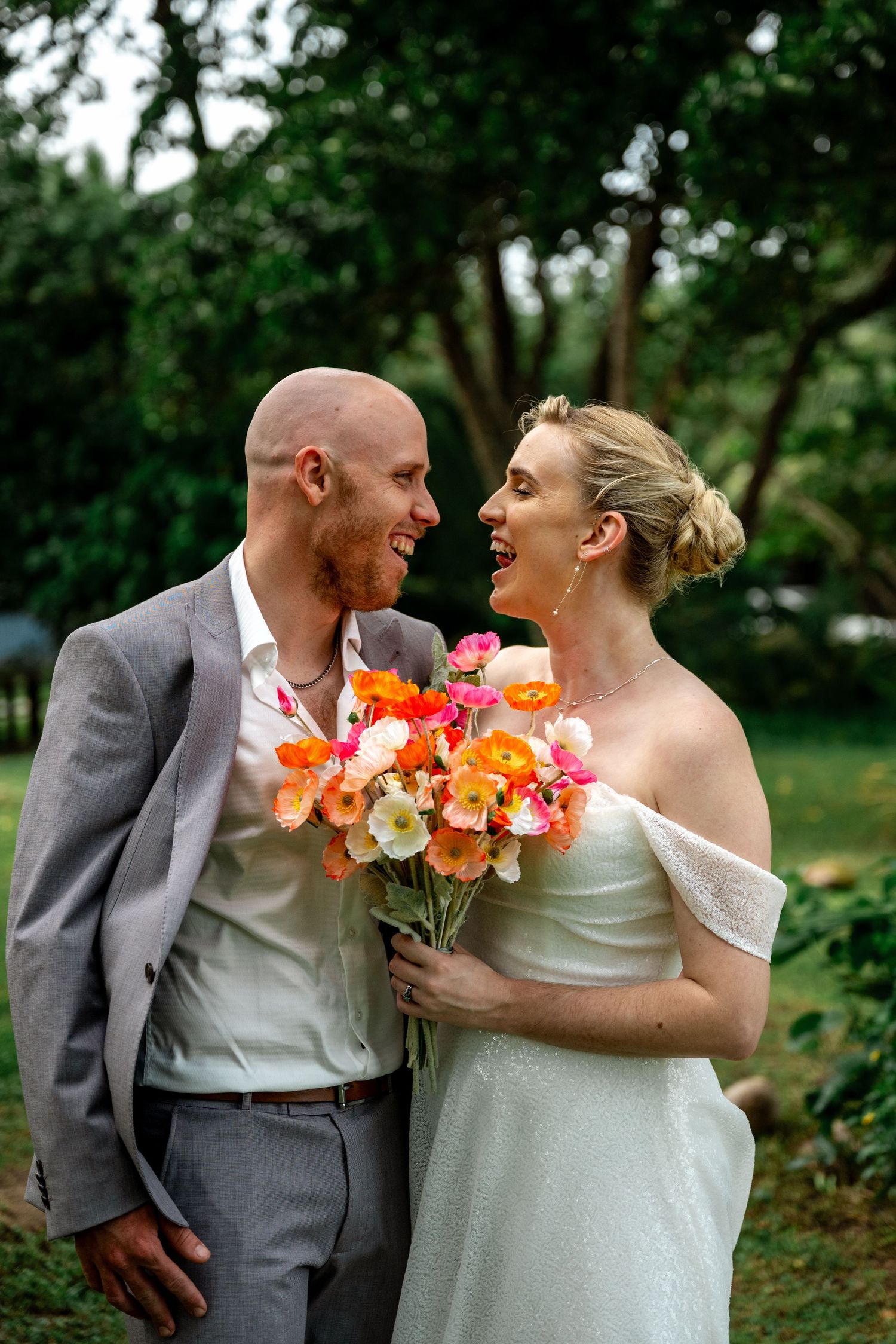 Laughing bride and groom sharing a joyful moment holding colorful orange and pink wedding bouquet in garden.
