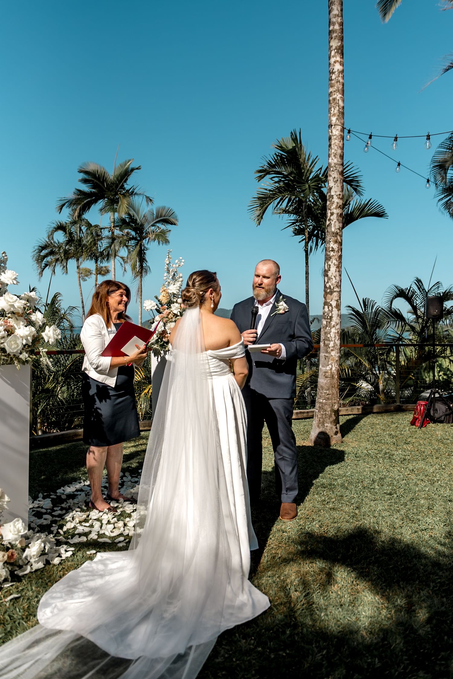 Bride and groom exchange vows at outdoor tropical wedding with white floral arch and palm trees backdrop.