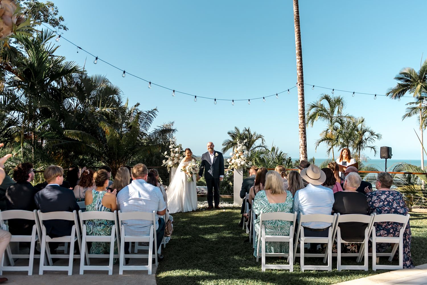 Guests seated at outdoor wedding ceremony with tropical palms and string lights overhead.