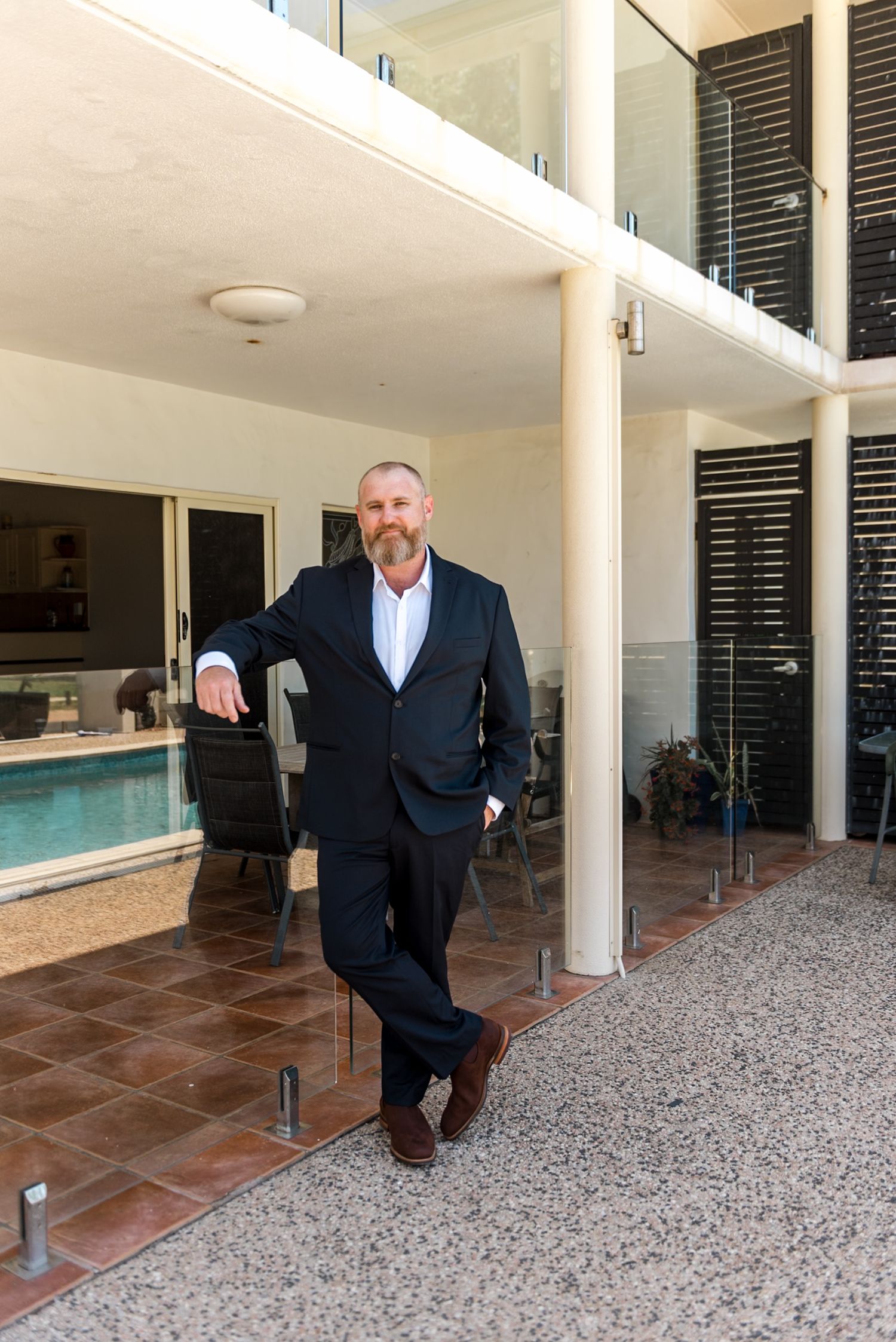 Middle-aged man in a dark suit leaning against a pillar near a pool area.
