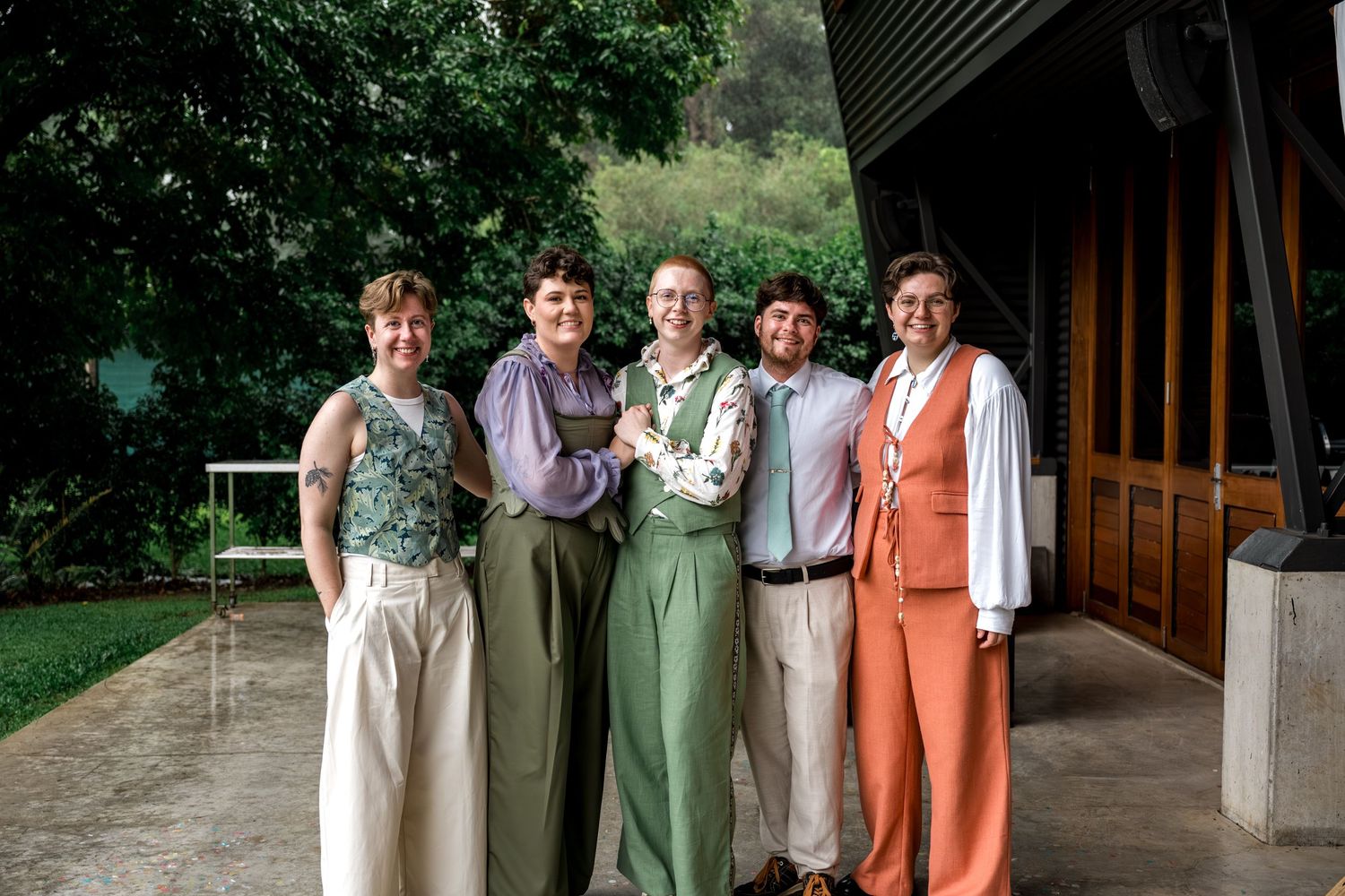 Group of five wedding guests smiling together outdoors in colorful formal attire.