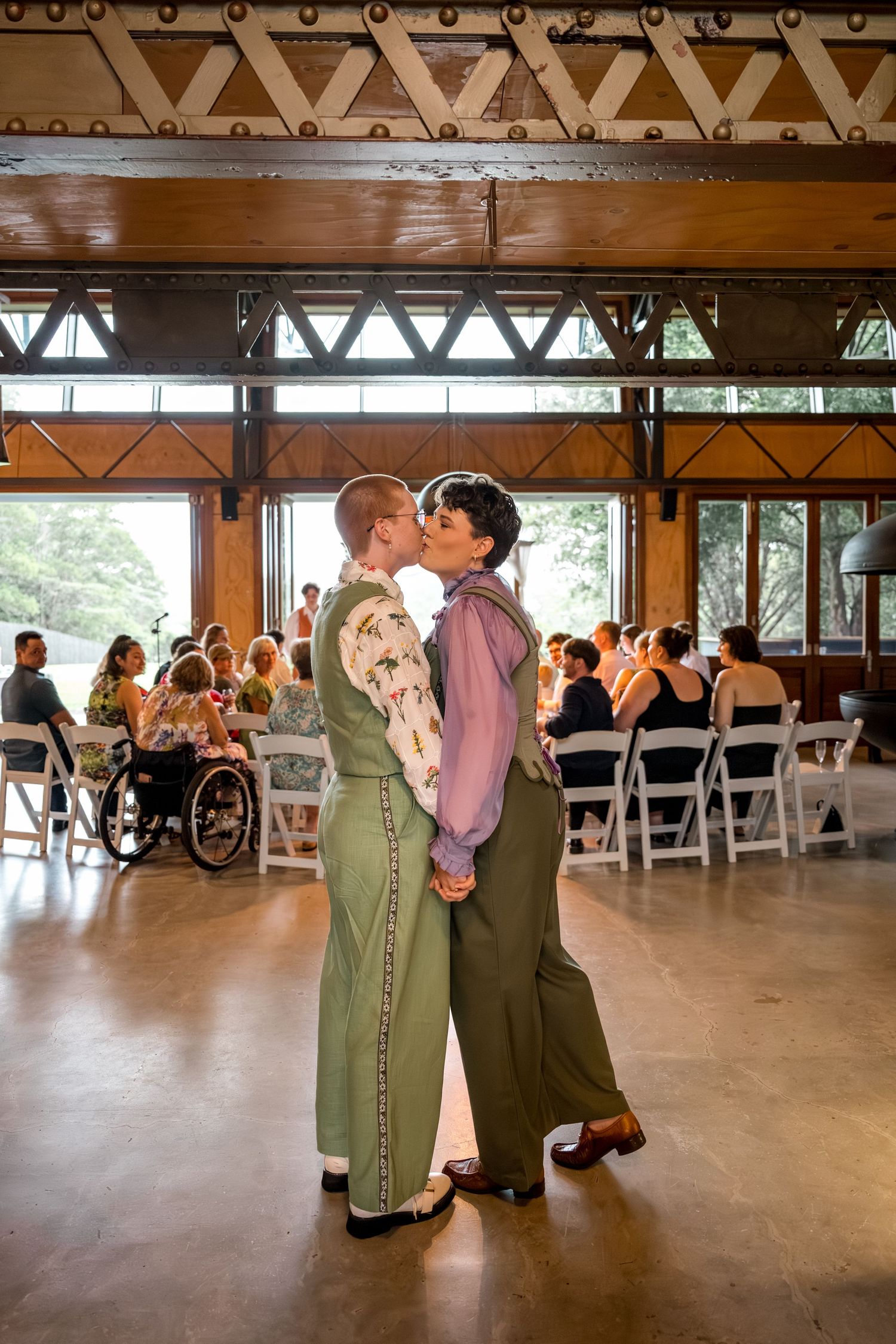 Two brides share a first kiss in green suits at their wedding ceremony in a rustic wooden venue.