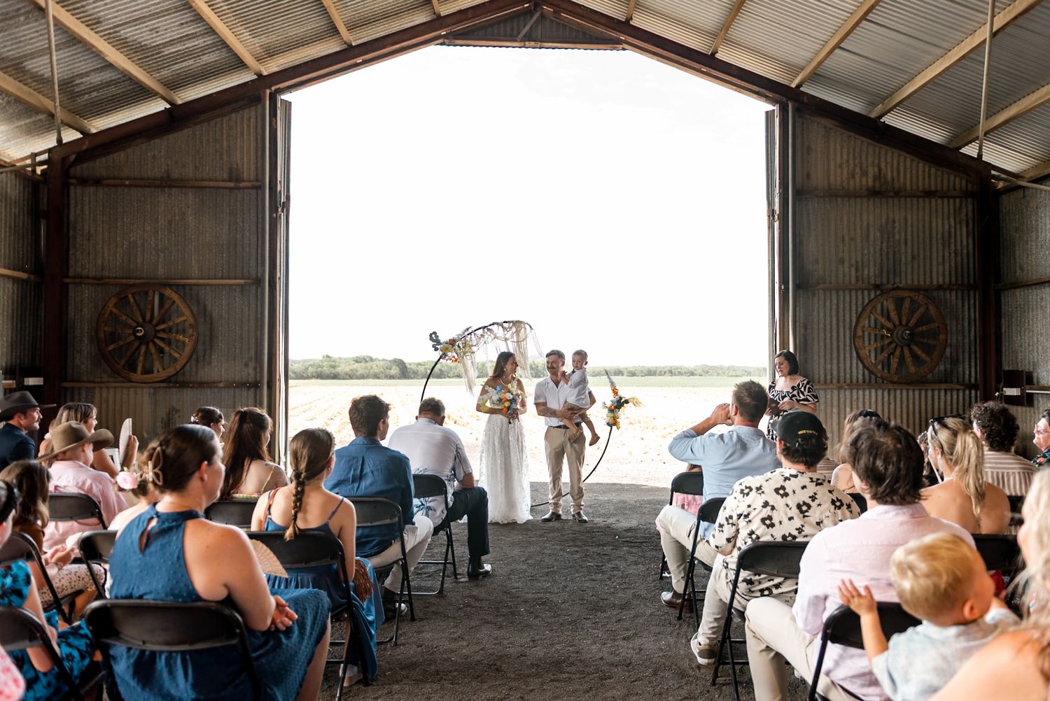 Rustic barn wedding ceremony with guests seated facing couple at boho macrame arch with open doors revealing countryside.