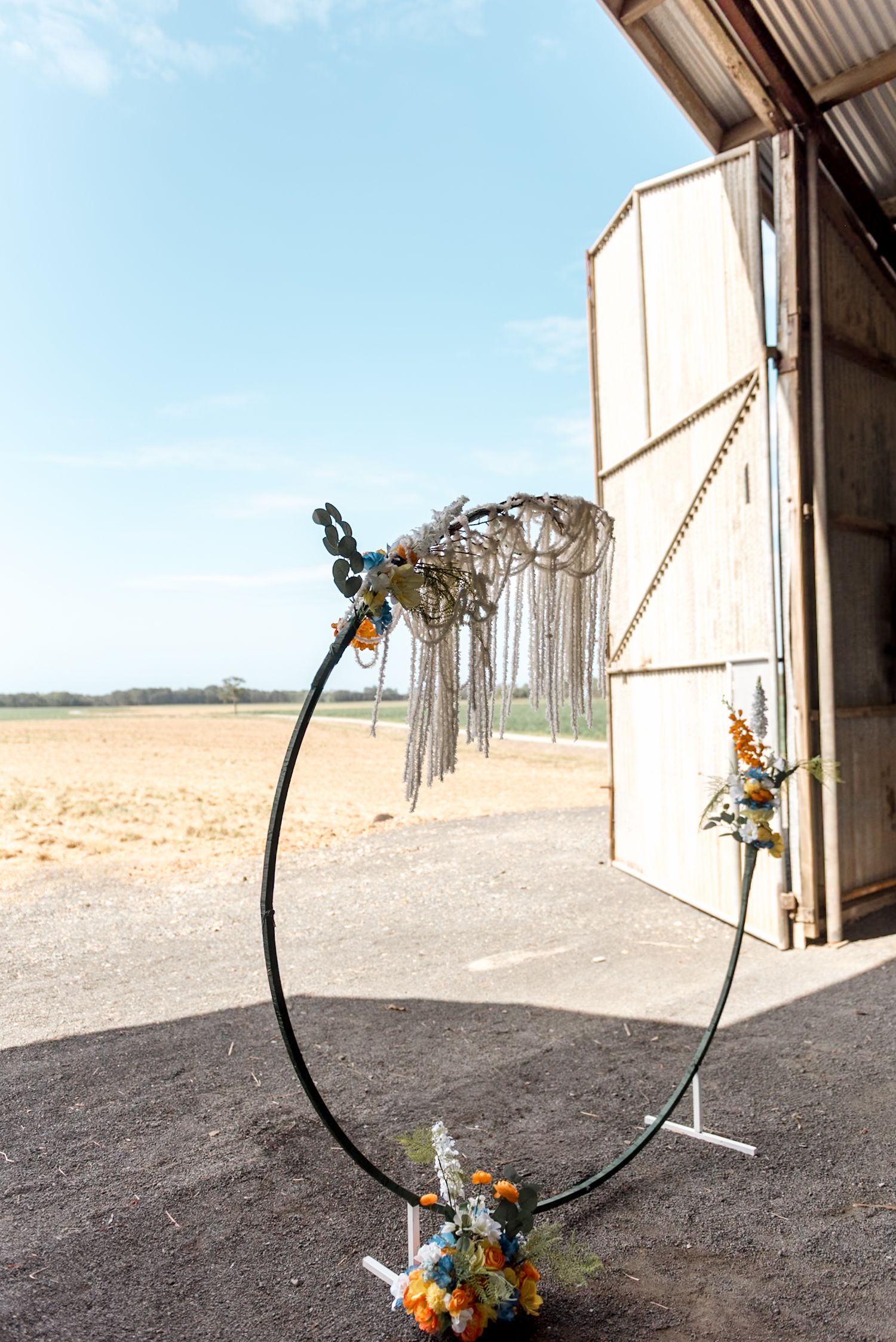 Circular metal wedding arch with macrame and floral decor outside a rustic barn.