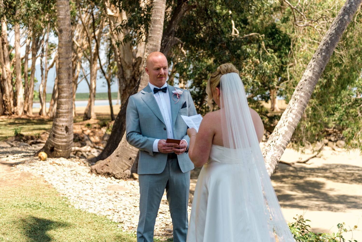 Smiling groom reads vows to bride at sunny tropical beach wedding under palm trees.