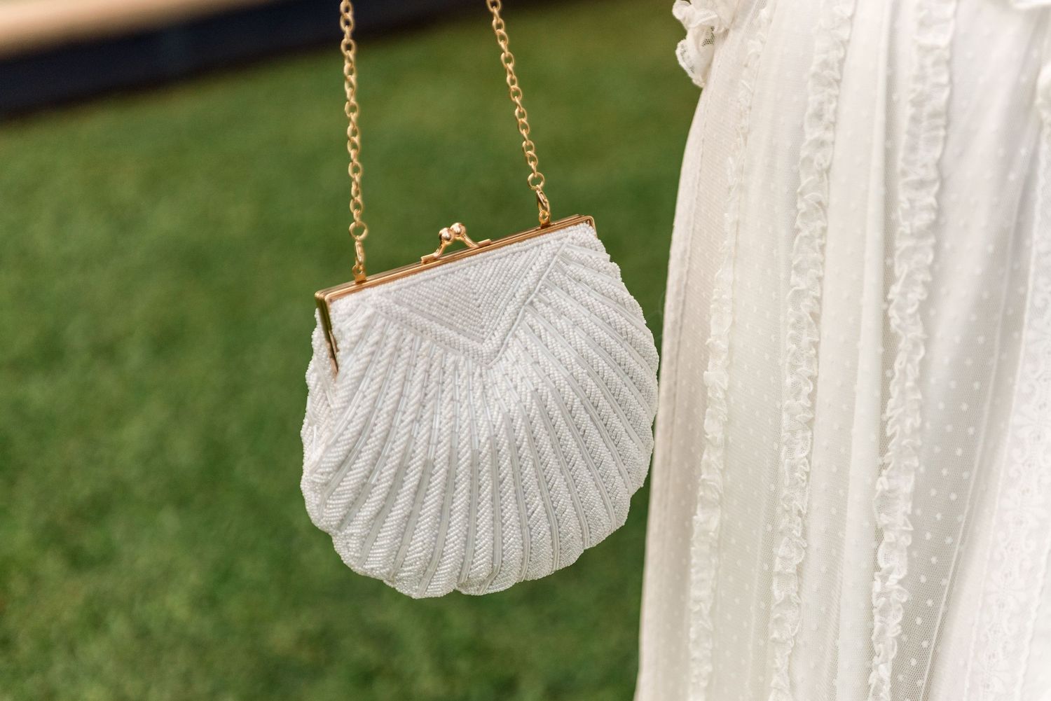 Elegant white beaded seashell-shaped bridal clutch bag with gold chain strap against a white dress.