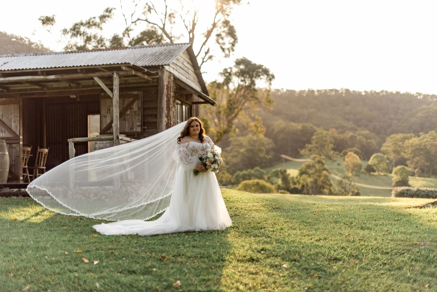 Bride in a flowing gown and veil poses outside a rustic barn at a golden hour farm wedding.