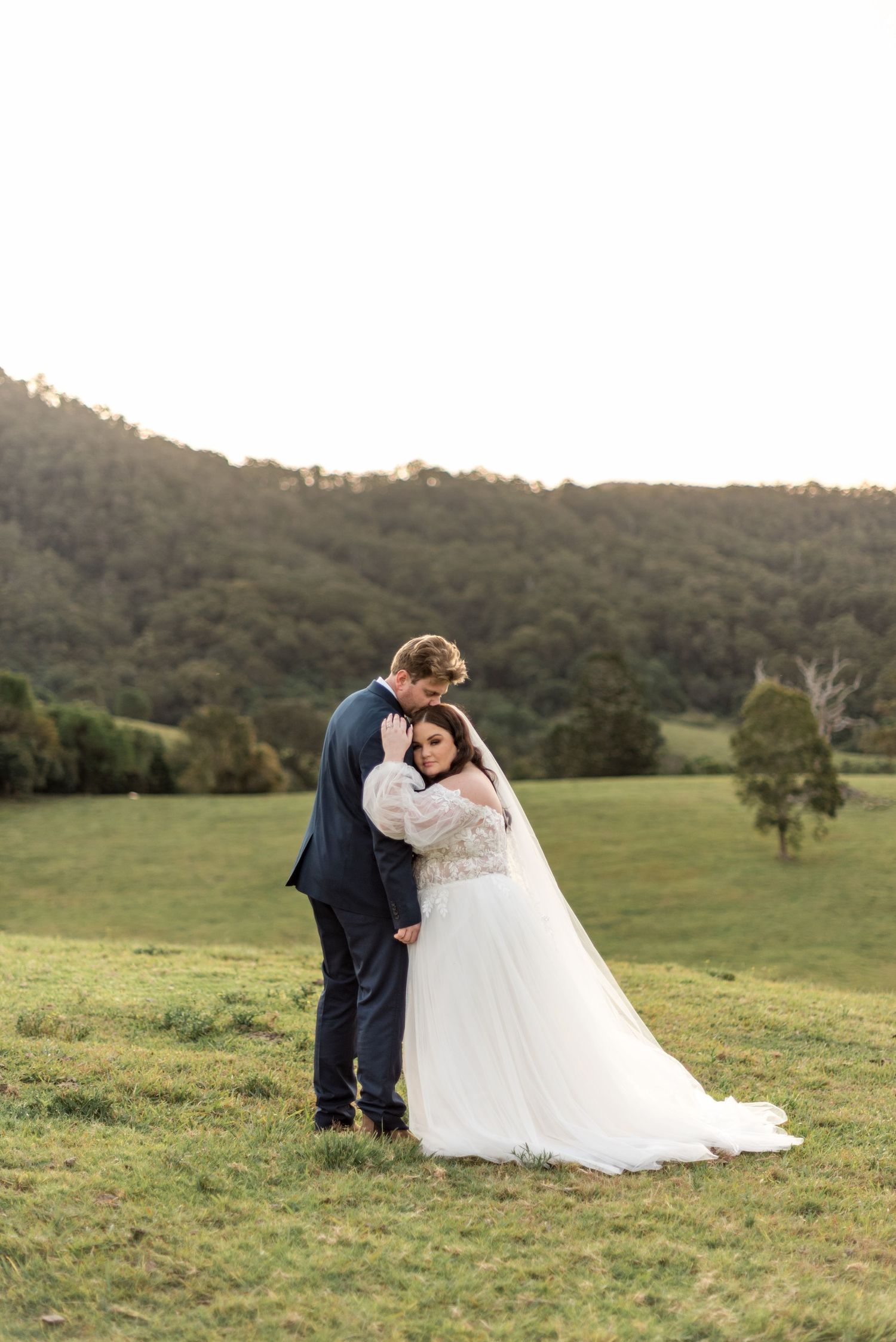 Groom in navy suit embracing bride in white gown against lush green mountain backdrop.