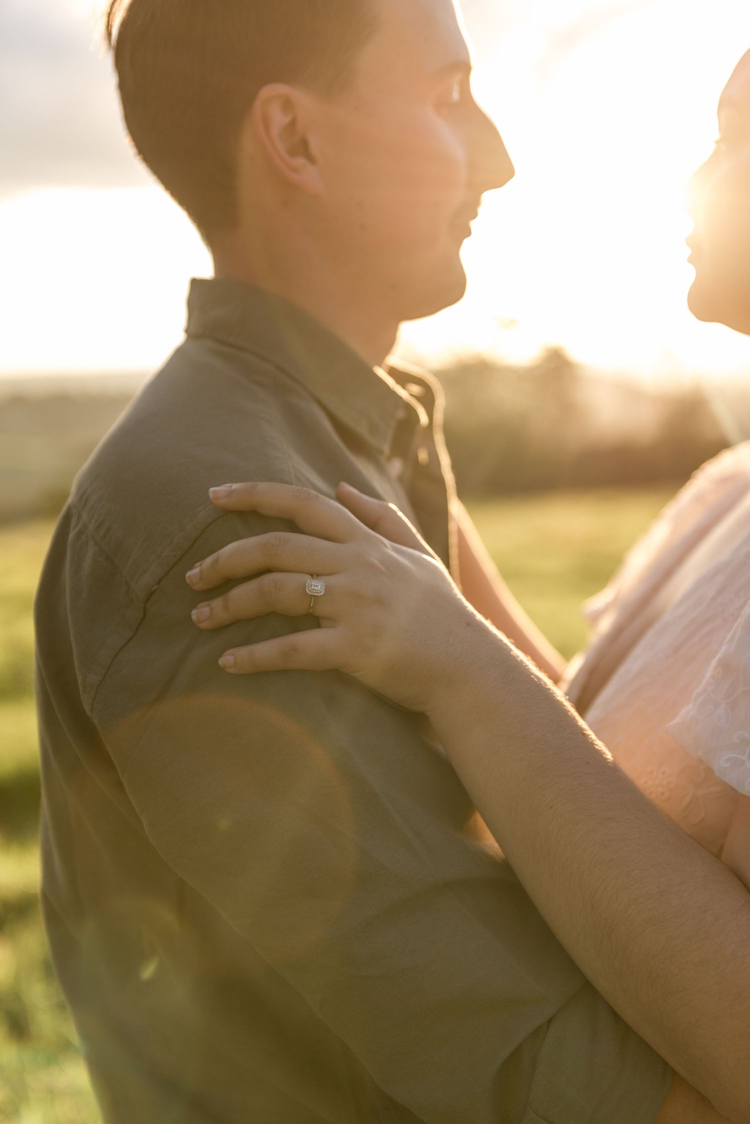 Close-up of a womans hand with engagement ring resting on mans shoulder at sunset.