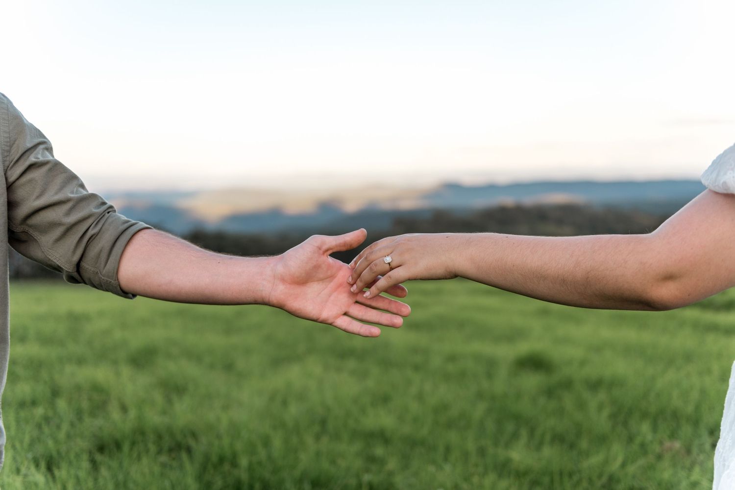 Couple reaching hands toward each other in green field at sunset, woman wearing diamond engagement ring.