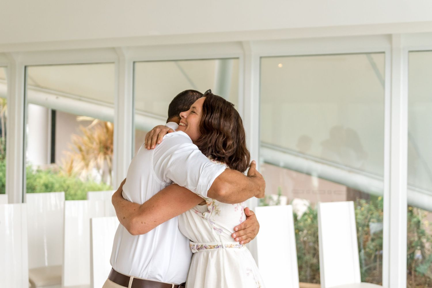 Two people sharing a warm embrace inside a bright, modern wedding venue with large windows.