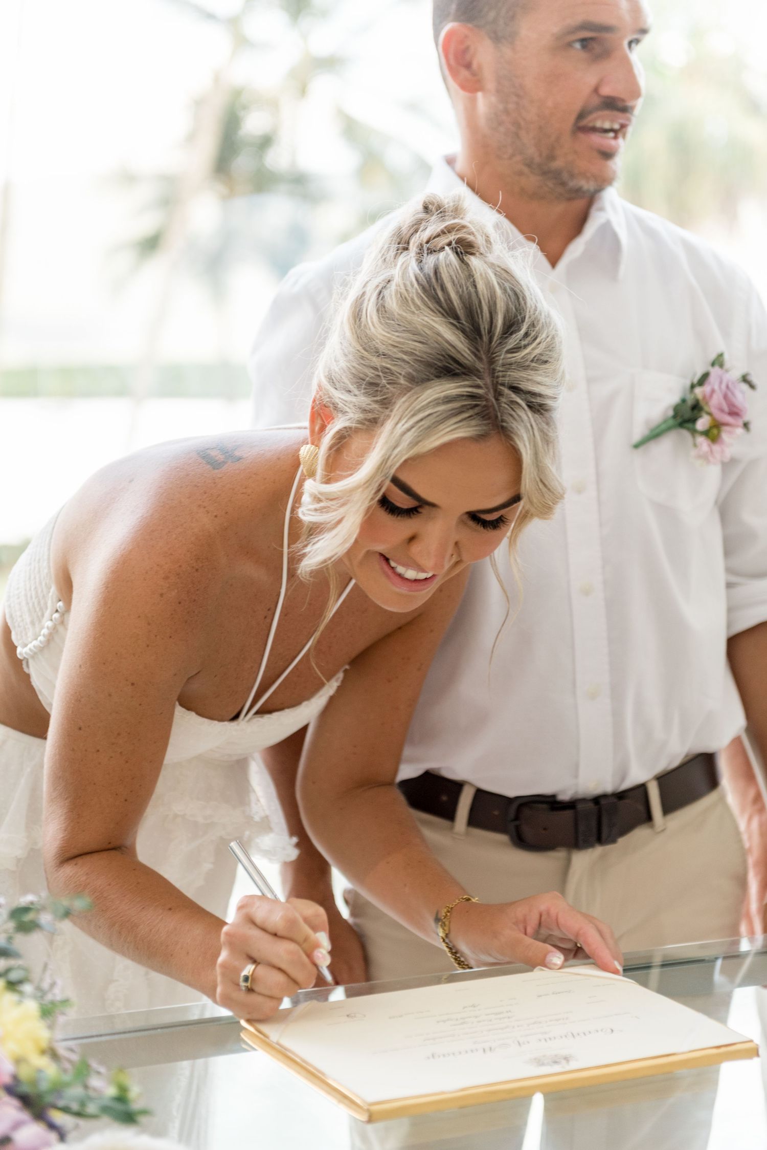 Smiling blonde bride signing wedding certificate while groom stands beside her at tropical venue.