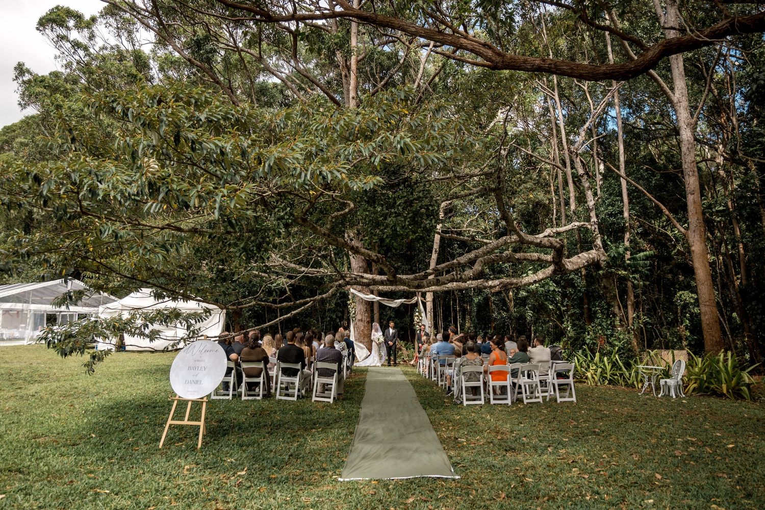 Outdoor wedding ceremony under large trees with white chairs and rustic arch in garden setting.