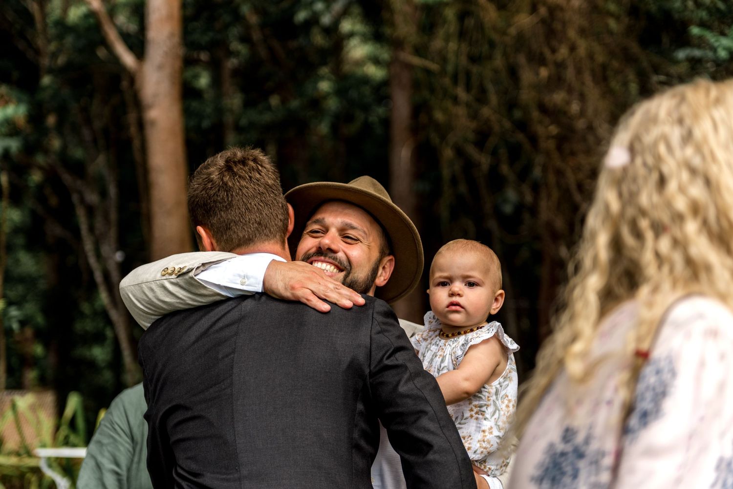 Smiling man in hat holding baby hugs groom after outdoor wedding ceremony.