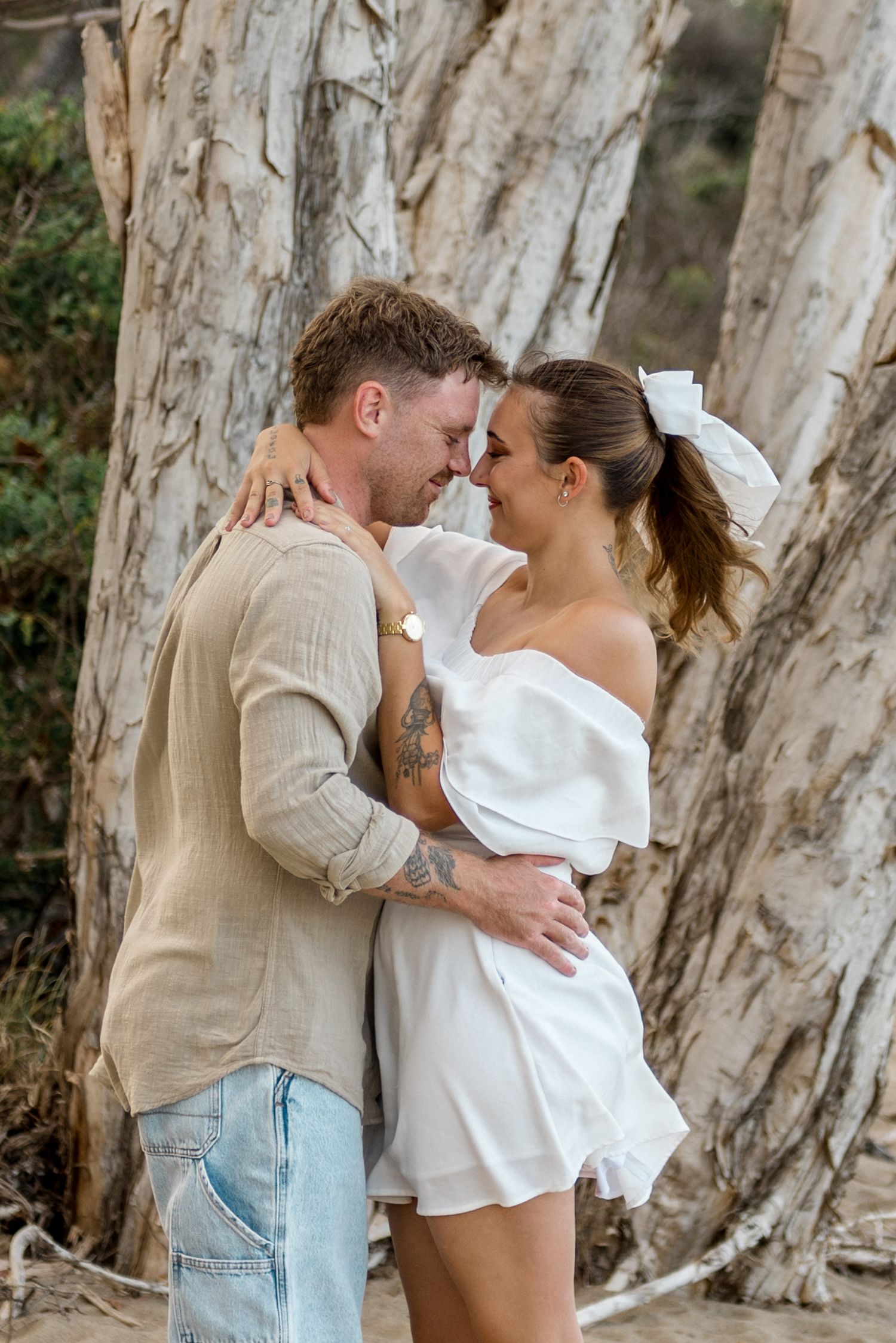 Couple in white dress and linen shirt embracing intimately against a large paperbark tree.