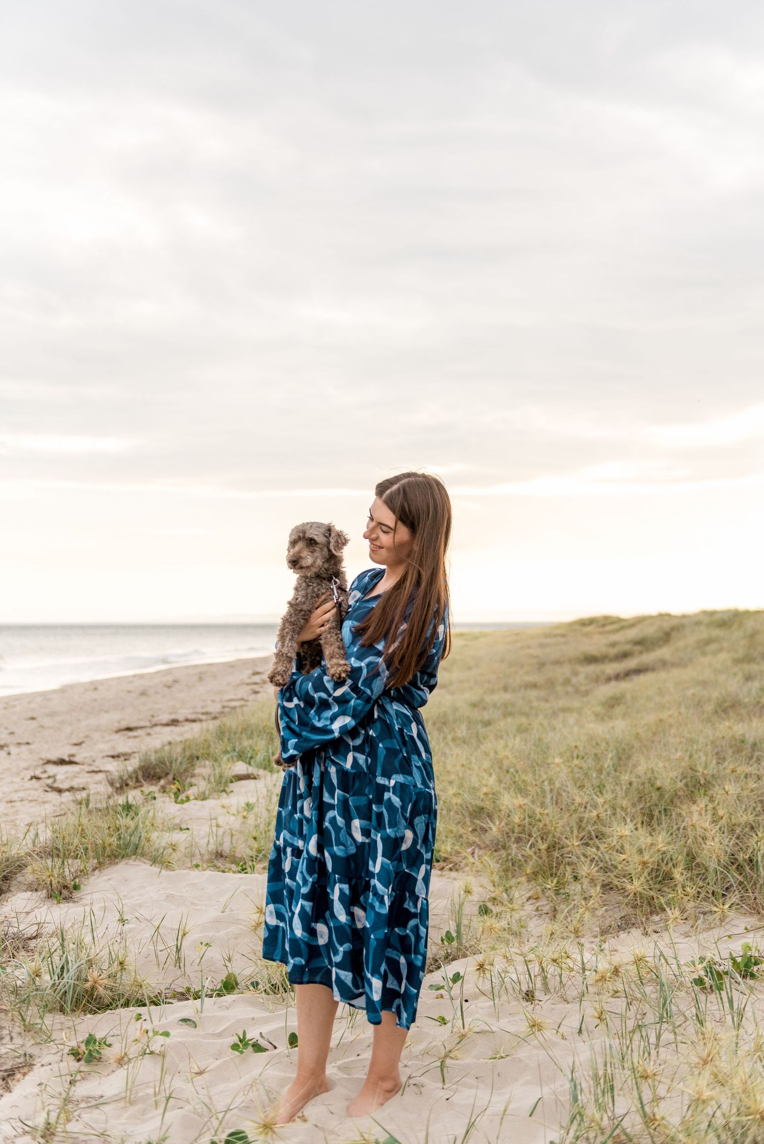 Woman in blue patterned dress holding small grey dog on sandy beach dunes at sunset.