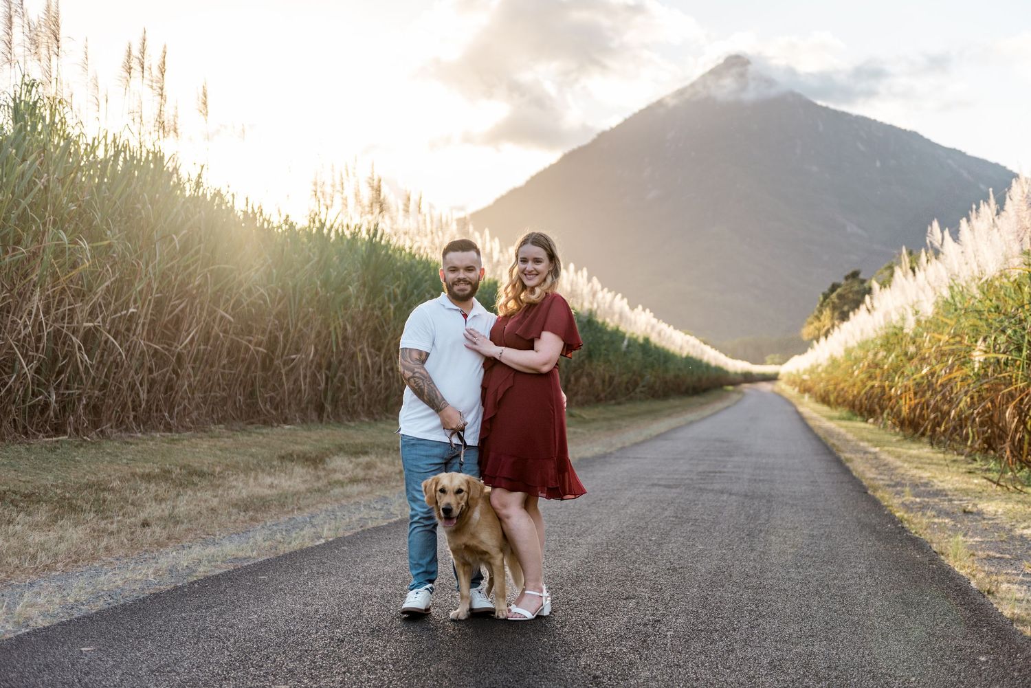Smiling couple with golden retriever standing on a rural road with mountain and sugarcane backdrop.