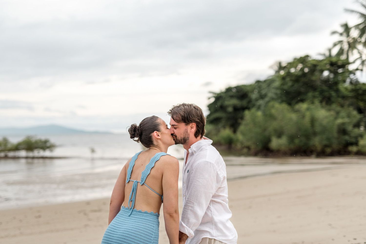 Couple sharing a kiss on a tropical sandy beach with palm trees and calm ocean waters in the background.