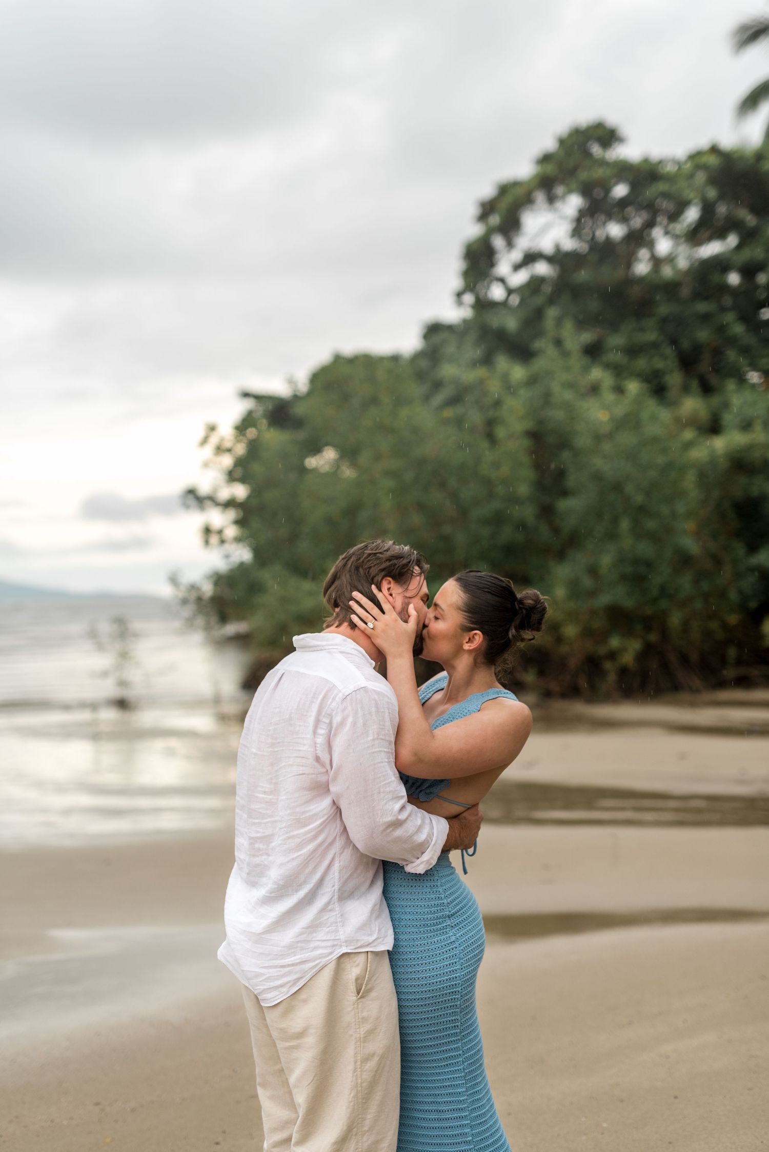 Lovers embracing and kissing on a sandy beach with ocean and tropical foliage behind them.