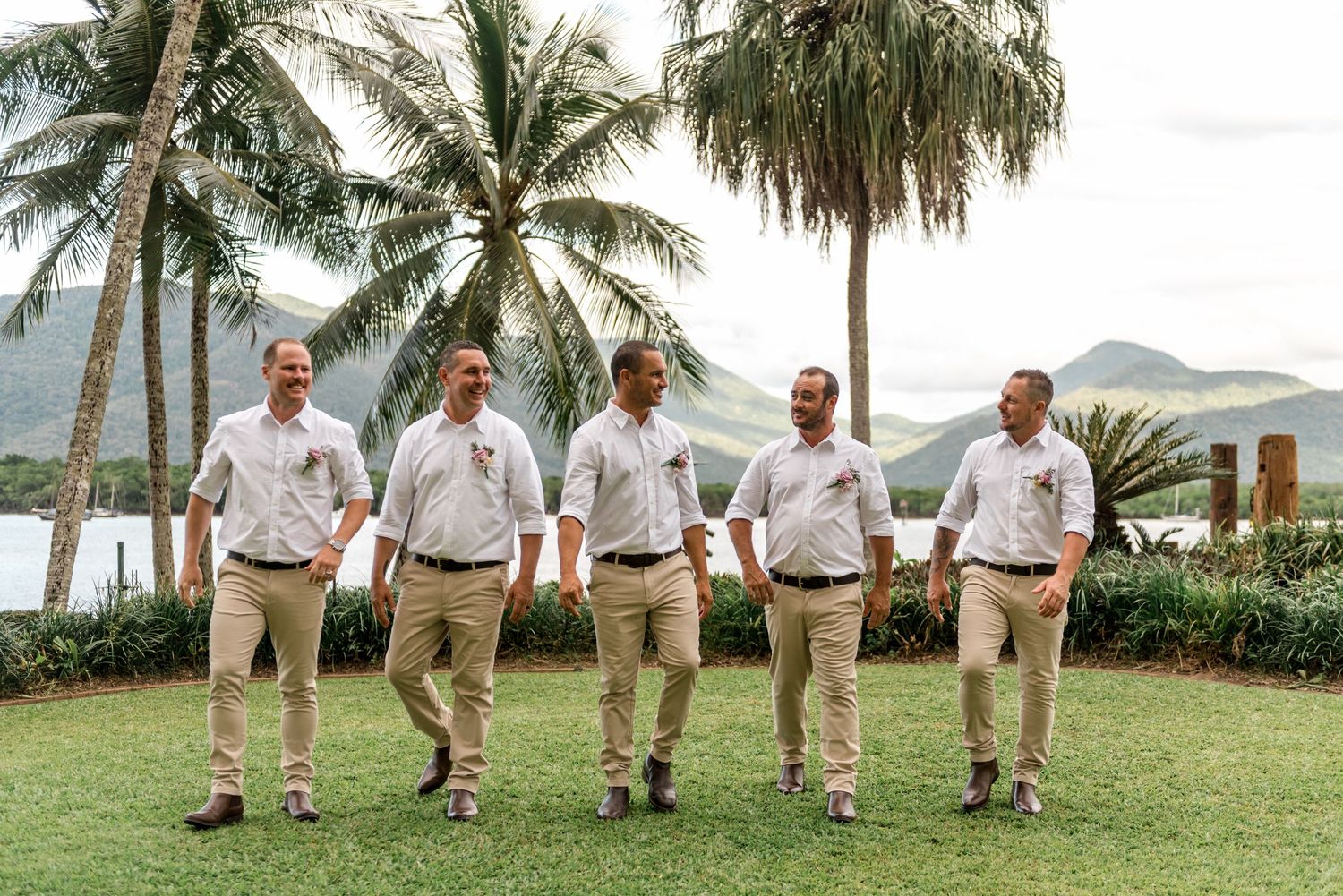 Five groomsmen in white shirts walk casually across lawn with palm trees and mountains behind.